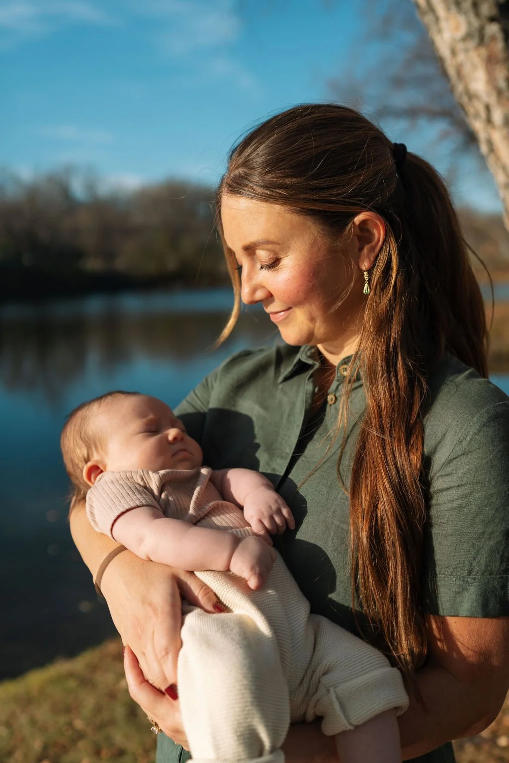 Dr. Andrea McNeer holding her sleeping baby outdoors near a body of water on a sunny day.