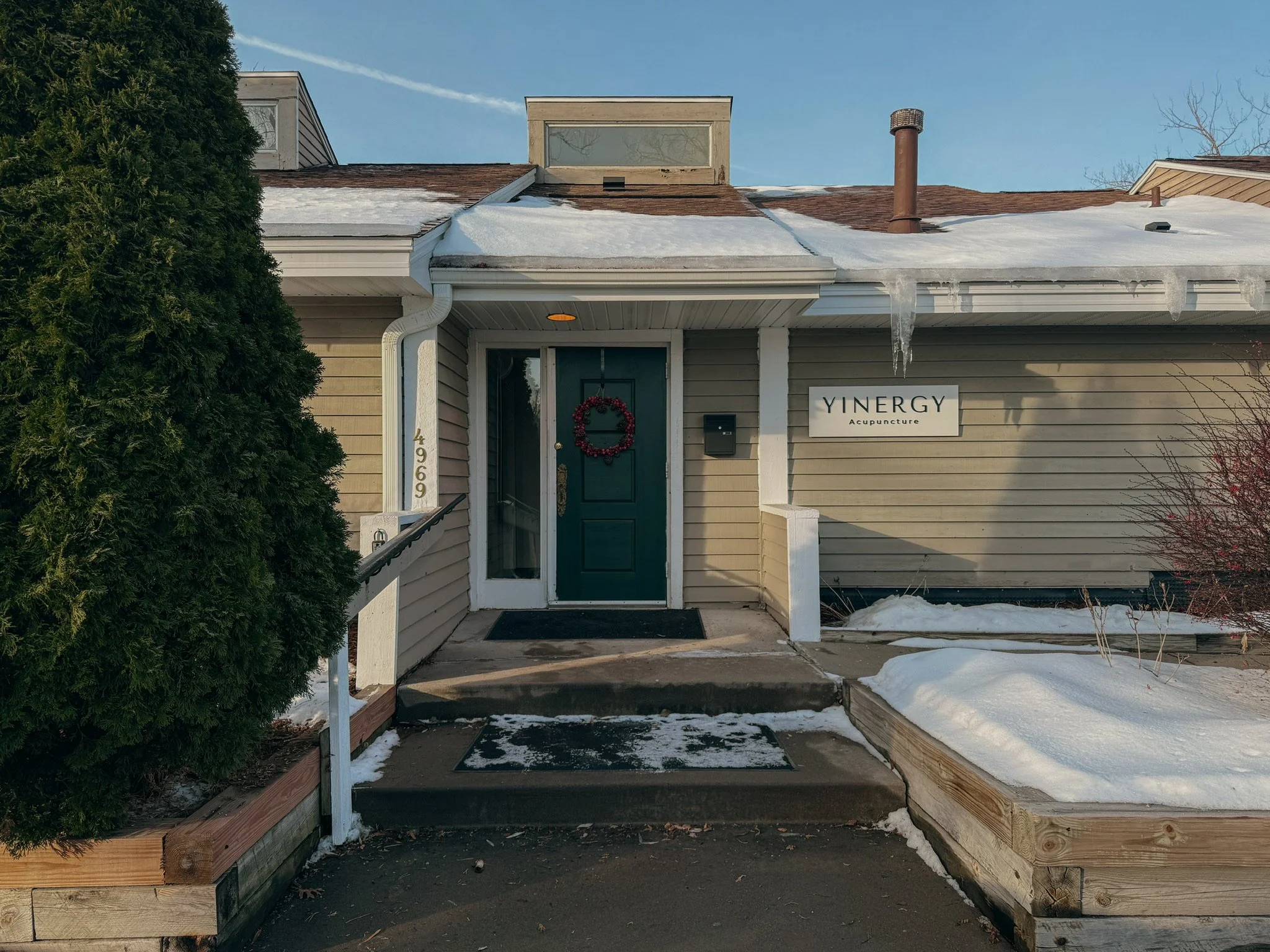 The front entrance of a Yinergy with snow on the ground and roof. The business has beige siding, a green door with a holiday wreath. There is a large bush on the left side and a small bare tree on the right.