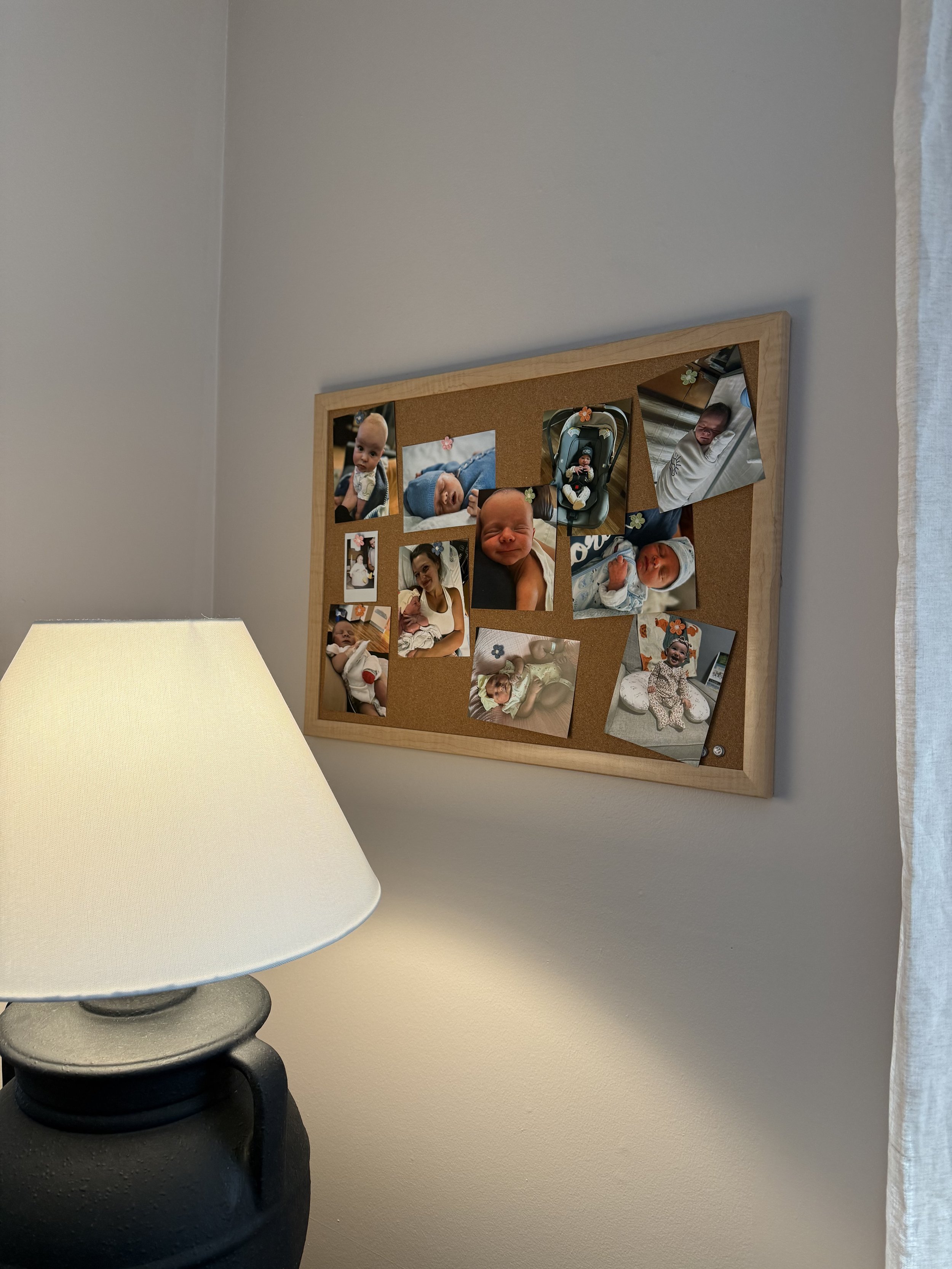 Cork bulletin board with multiple photos of babies and women, in a room with a beige wall and a lamp. This is Yinergys Baby Wall