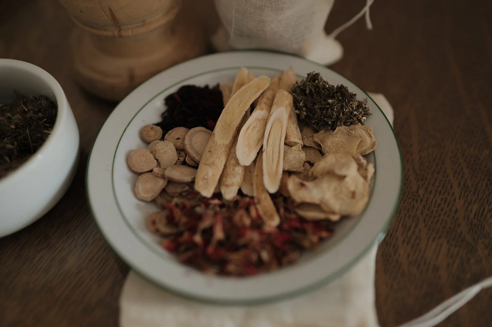 A white plate with various Traditional Chinese dried herbs and roots on a wooden table, with other containers of herbs nearby.