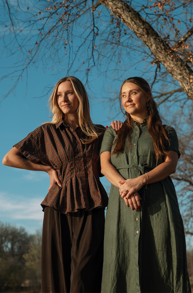 Dr. Hannah Zeleznikar and Dr. Andrea McNeer standing outdoors under a leafless tree against a blue sky, with one woman resting her hand on the other's shoulder, both looking to the side.