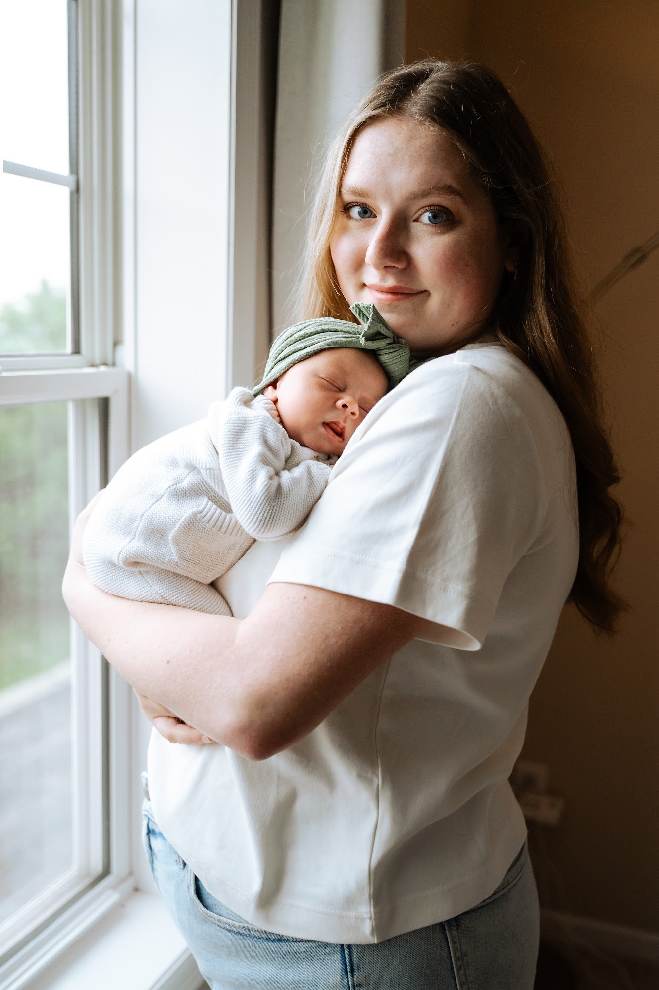 A young woman with long brown hair holding a sleeping baby near a window, both in casual clothing.