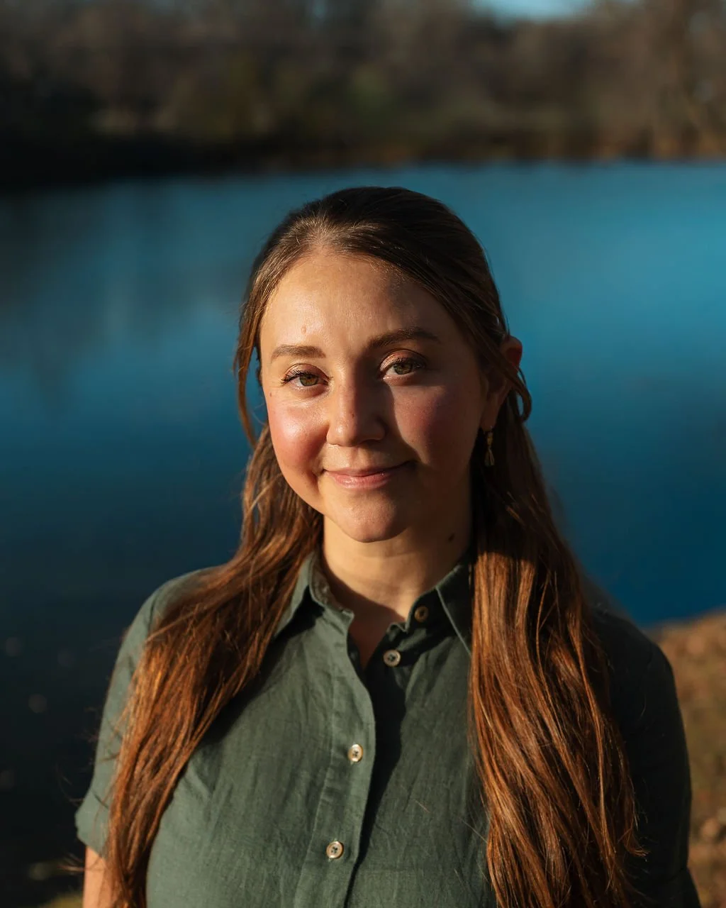 Dr. Andrea McNeer, A woman with long brown hair and a green button-up shirt smiling outdoors near a body of water during sunset or sunrise.