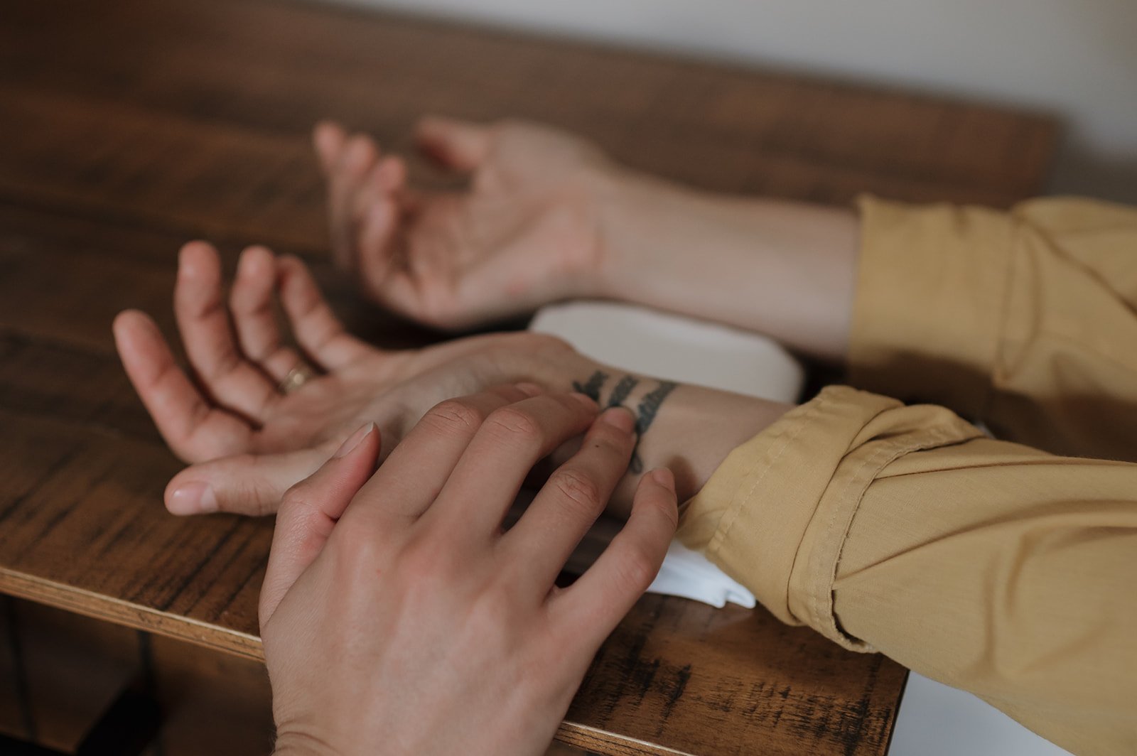Close-up of two hands, one with a tattoo, resting on a wooden surface; one hand is taking the pulse of the other. The person with the tattoo is wearing a yellow shirt, and the image focuses on the hands and forearms.