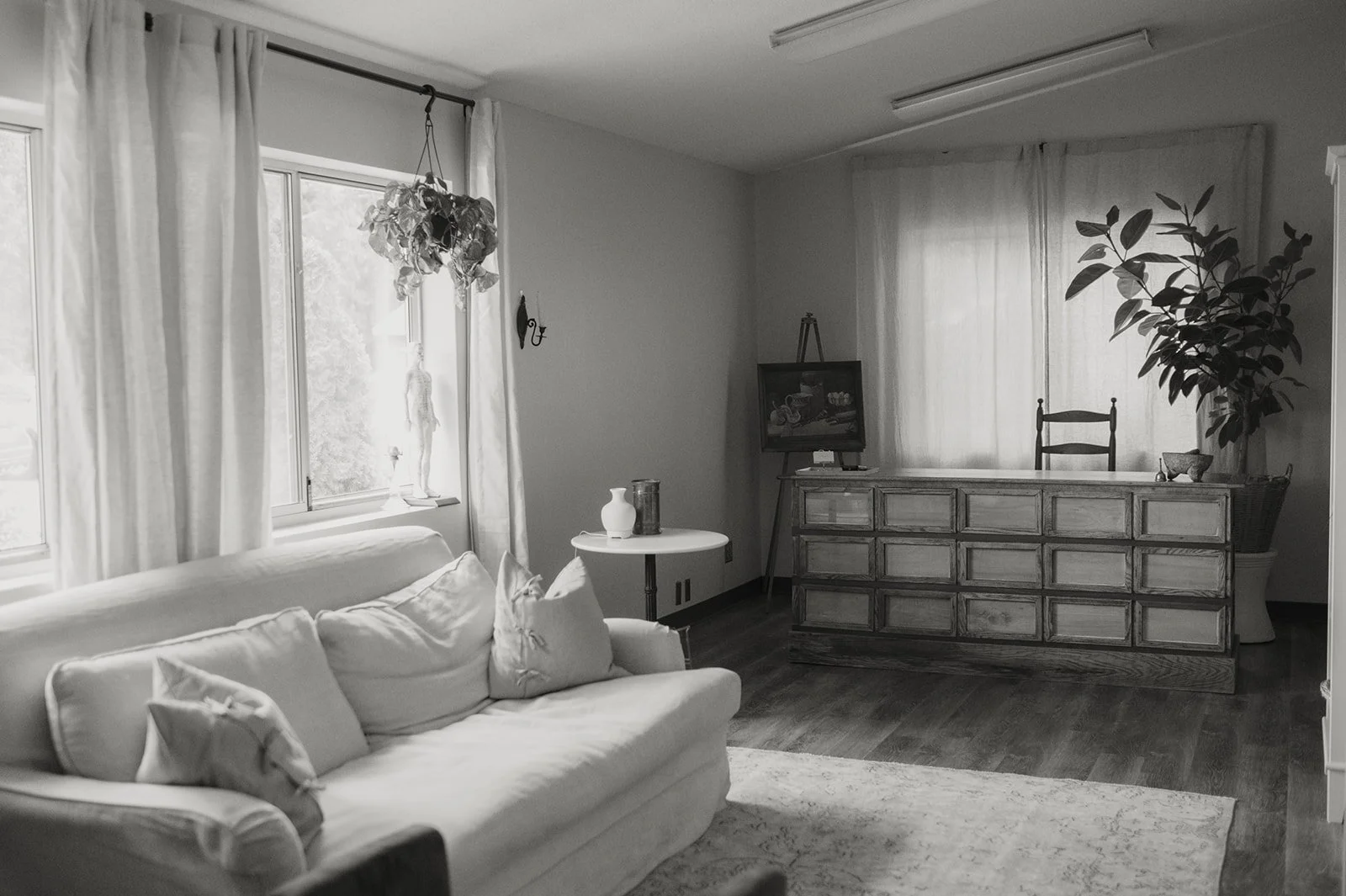 Yinergys Lobby, a room with a sofa, cushions, a window with curtains, a small table with vases, a chest of drawers, a chair, a large plant, and a painting on an easel.