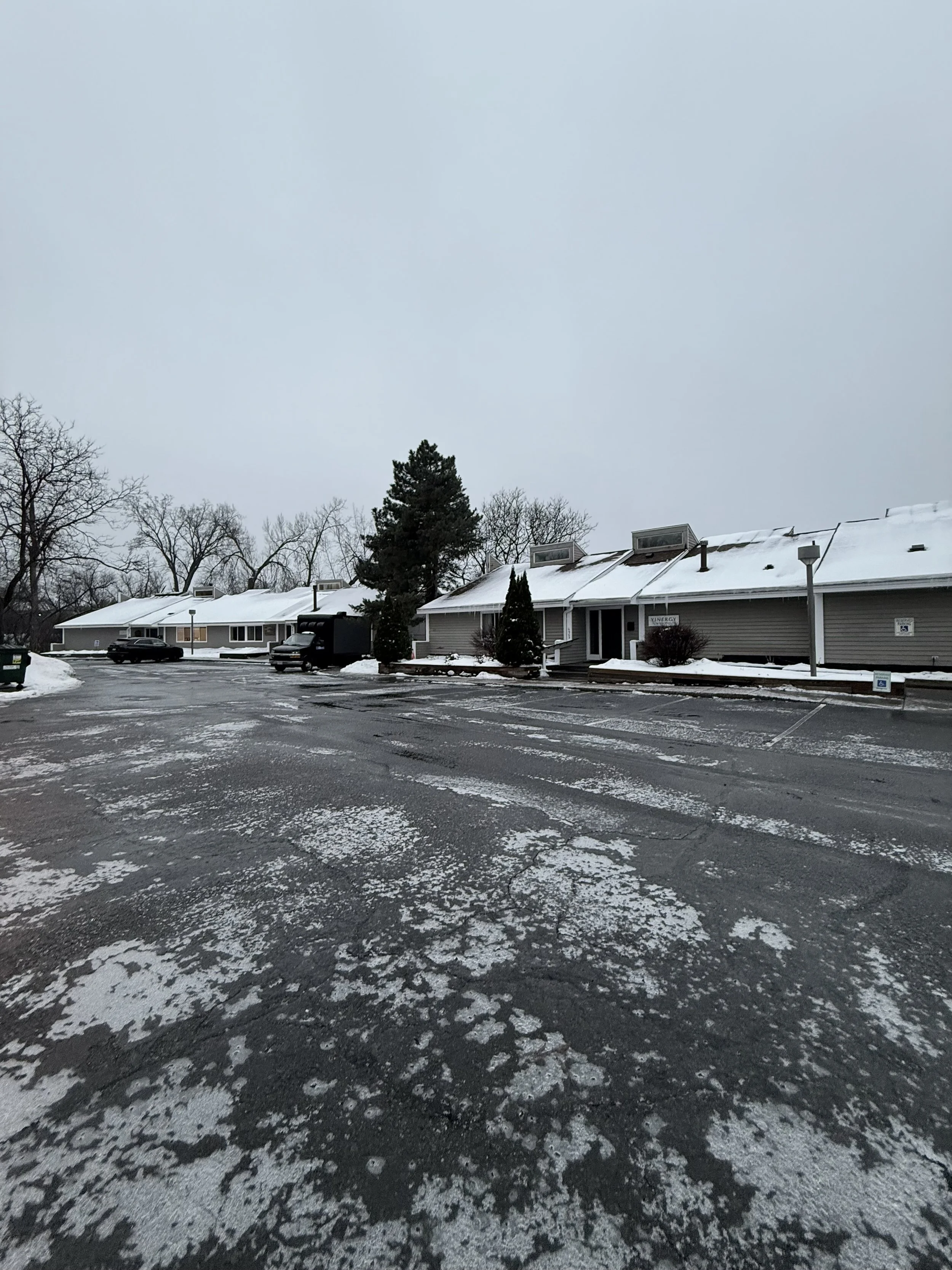 Cold, snowy parking lot outside a row of beige business buildings with snow-covered roofs and trees in the background, overcast sky.