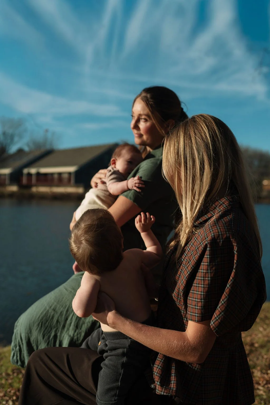 Dr. Hannah Zeleznikar and Dr. Andrea McNeer sitting outdoors by a lake, holding their young children. The scene is during the daytime, with houses and trees in the background under a blue sky in the back yard of Yinergy Acupuncture.