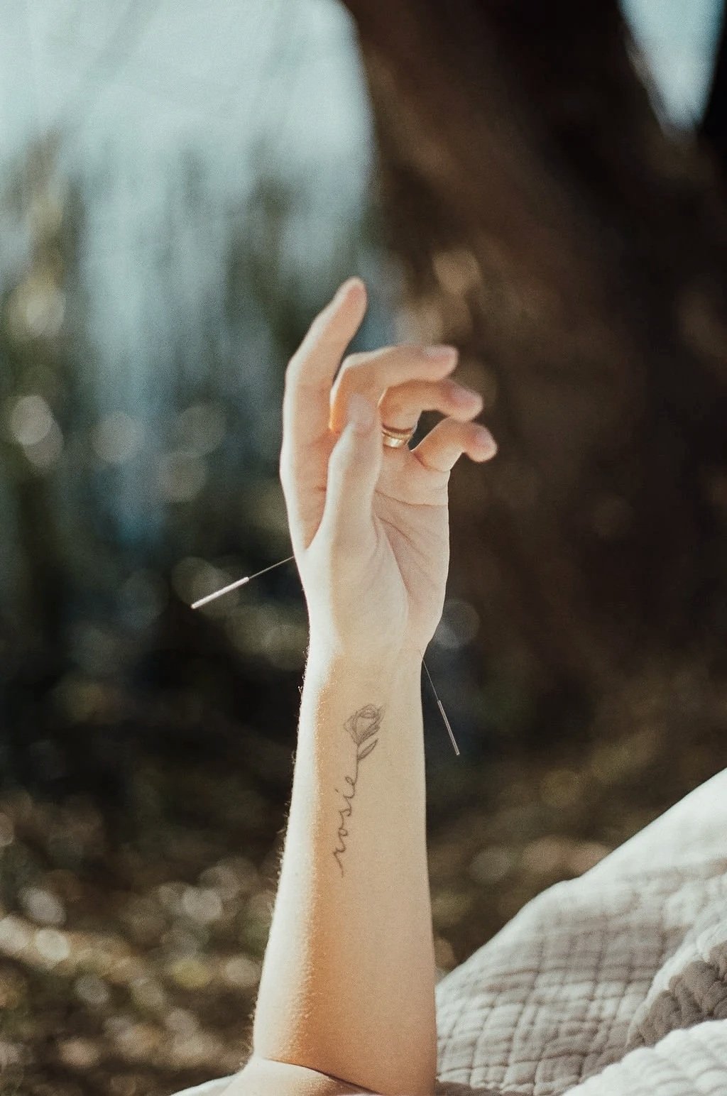 A close-up of a woman's hand with acupuncture needles in it, with a tattoo of a face outline and the word 'smile' on her forearm. The background is blurred.