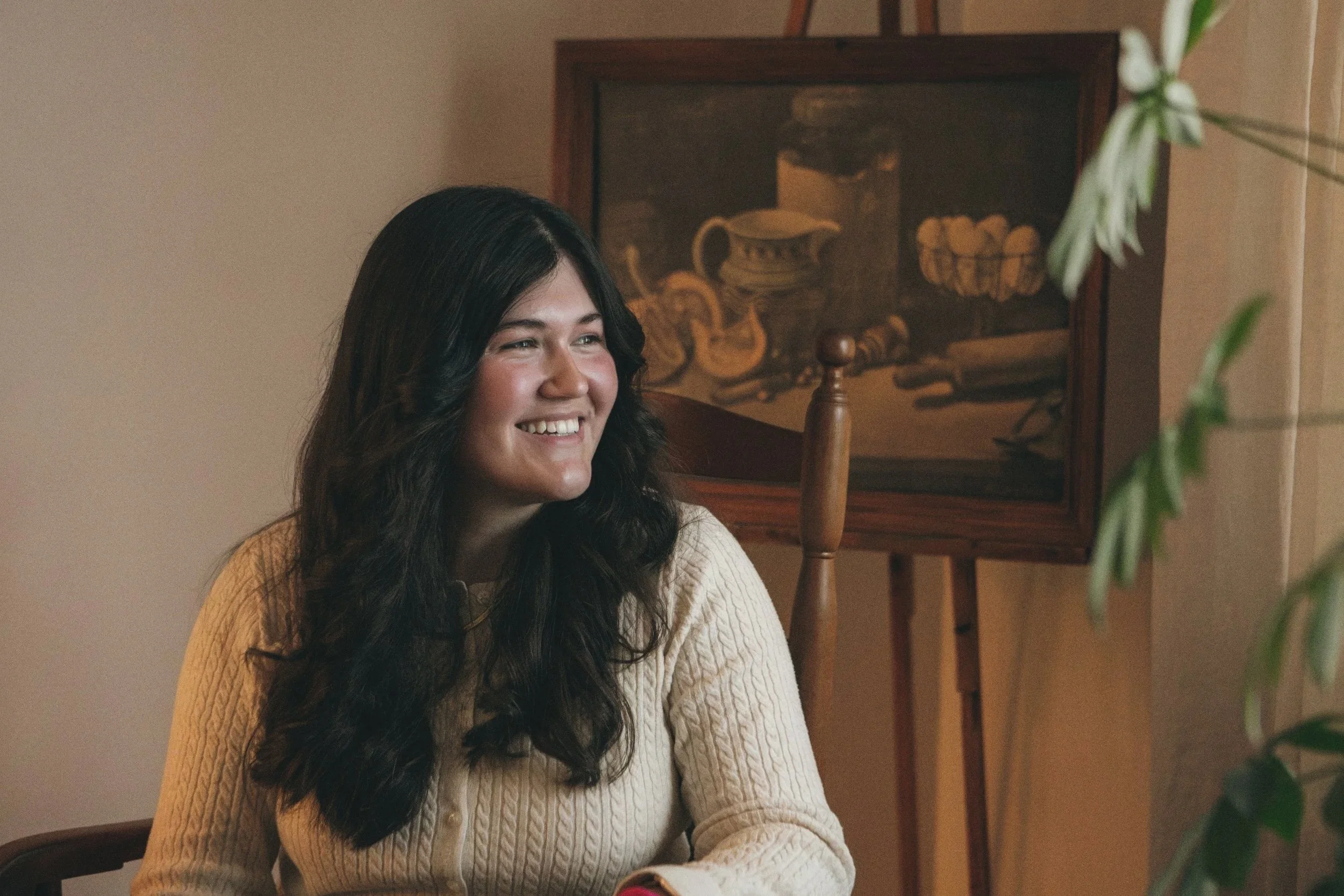 Lilly Anderson, Yinergys Office Manager and Media Manager, A young woman with long dark hair smiling while sitting indoors near a painting and a window with curtain, with a green plant in the foreground.