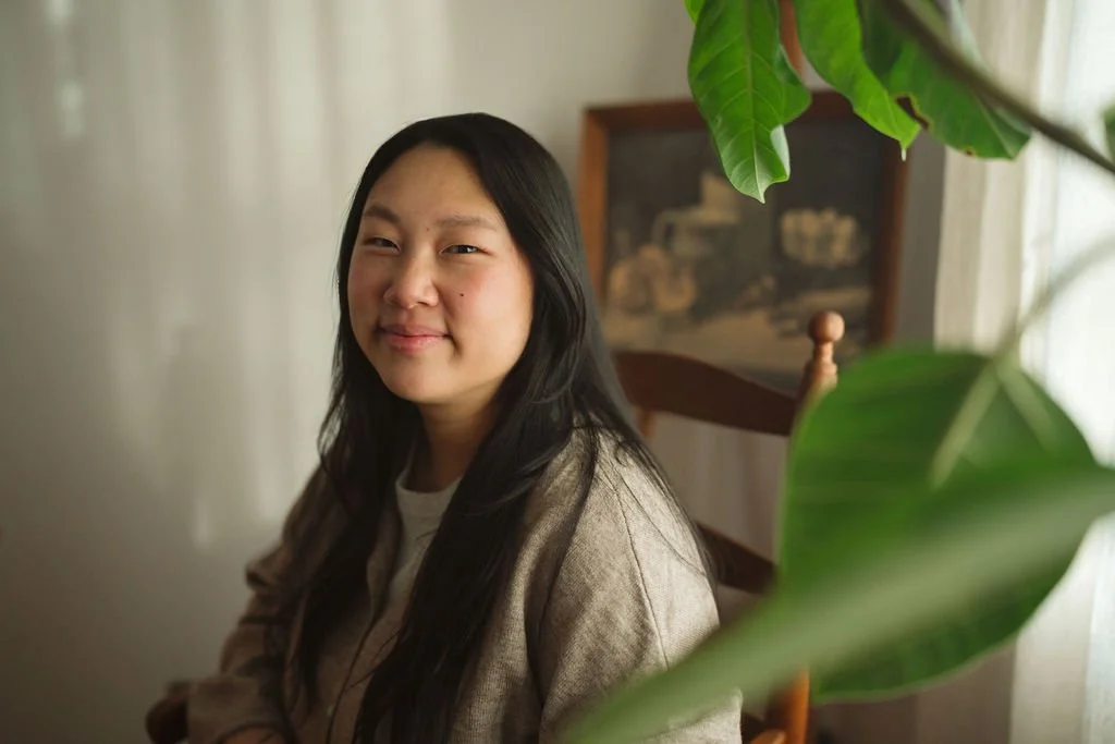 Nicole Mun, Receptionist, Acupuncture Student, A woman with long black hair smiling indoors, with green leaves in the foreground and a framed picture on a wooden shelf in the background.