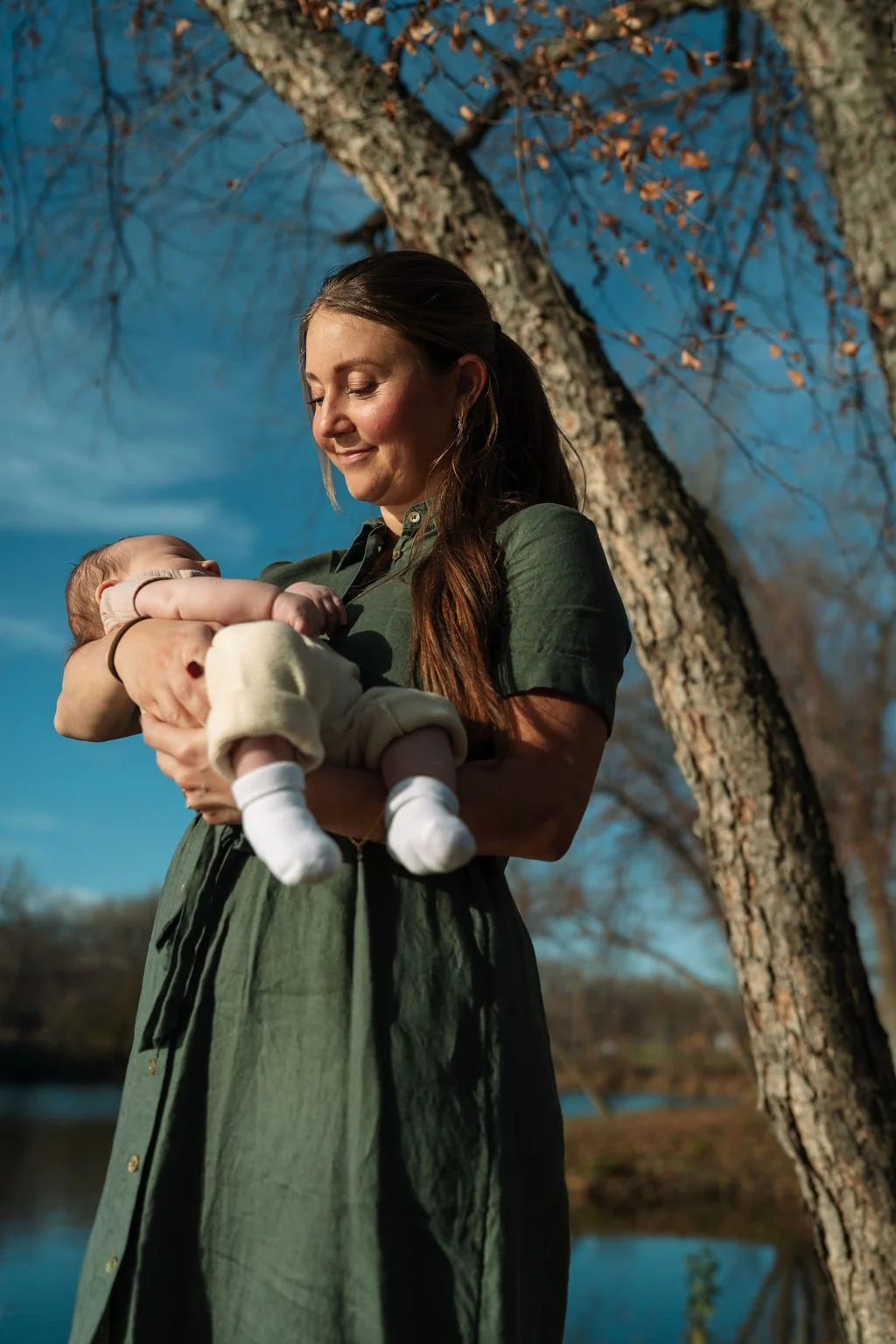 Dr. Andrea McNeer, A woman holding a baby outdoors next to a large tree, with a river in the background and a clear blue sky.