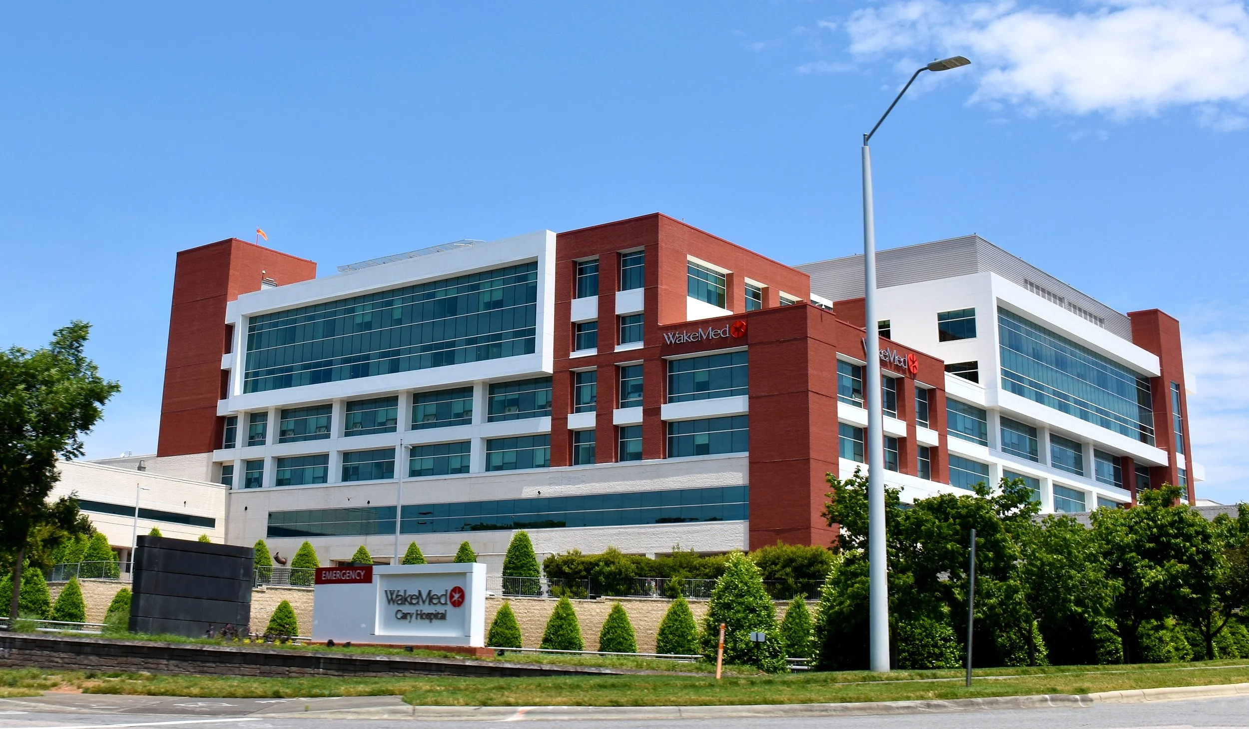 Exterior view of WakeMed Cary Hospital with a sign in the foreground, trees, a lamppost, and a blue sky with clouds.