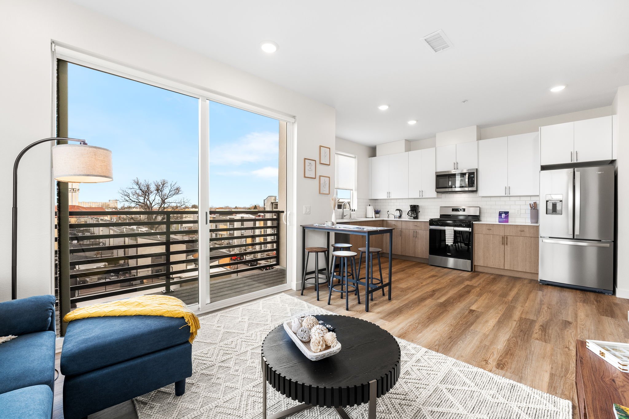 Open-concept living room and kitchen with large sliding glass door leading to balcony, white cabinets, stainless steel appliances, small dining table with stools, and wood flooring.