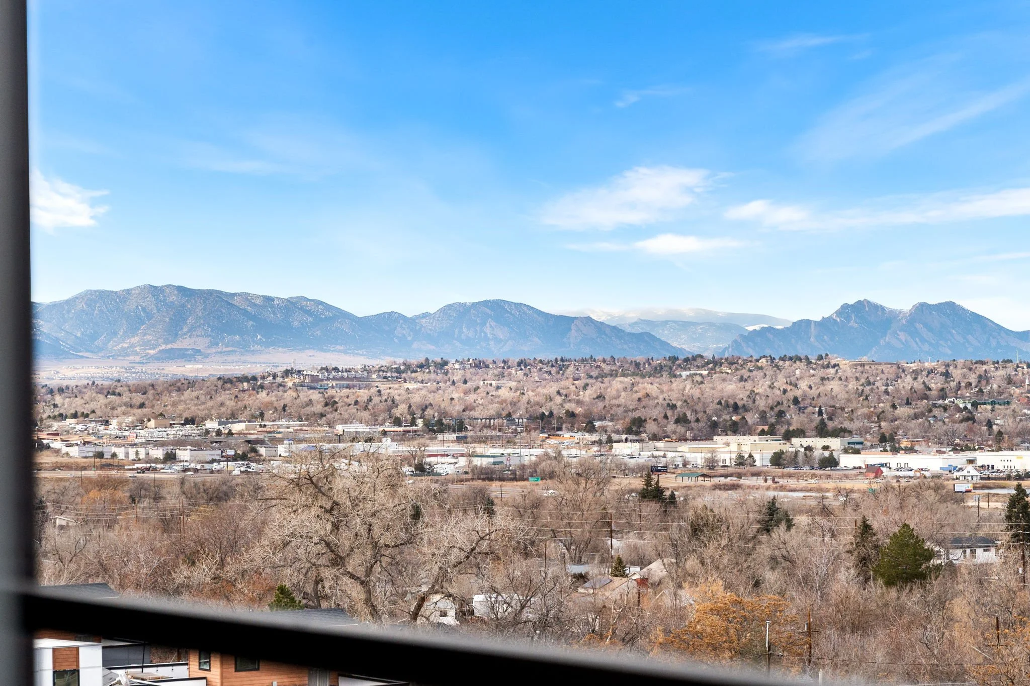 View of distant mountains, clear blue sky, a mix of leafless trees and evergreen trees in the foreground, suburban area with houses and commercial buildings in the middle ground.