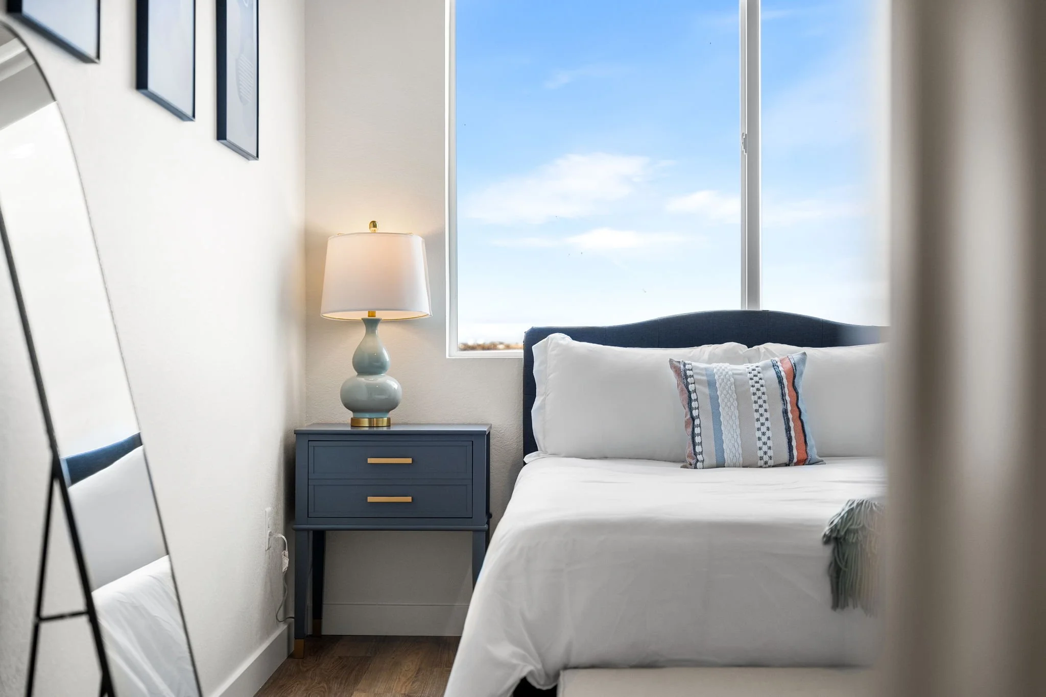 A modern bedroom with a bed, a blue nightstand, a lamp, and a window showing a blue sky.