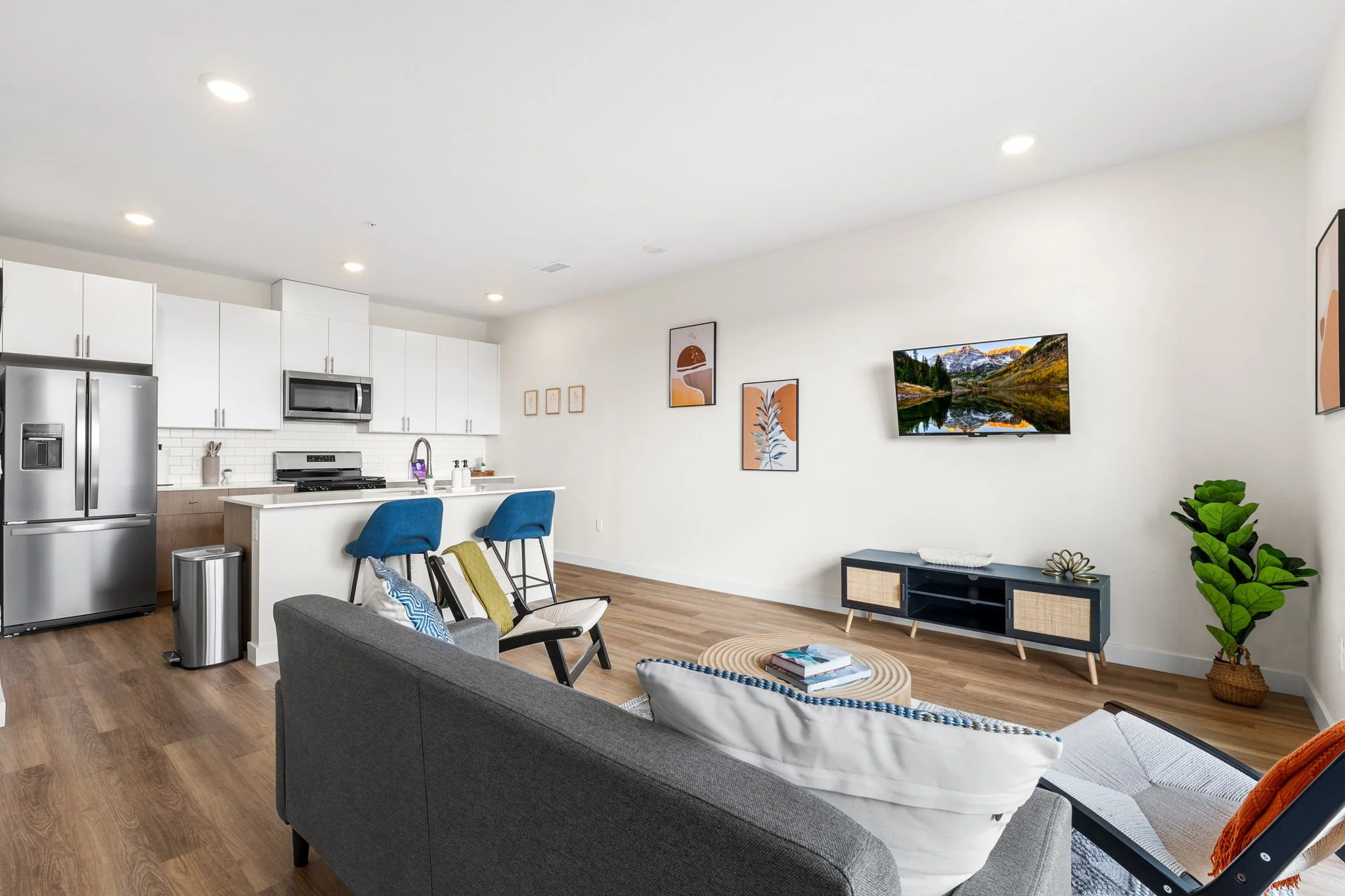Open-concept living room and kitchen with white cabinets, stainless steel refrigerator, microwave, and stove; blue bar stools at a white kitchen island; gray sofa with pillows; black TV stand with decorative items; wall art and a flat-screen TV on wh