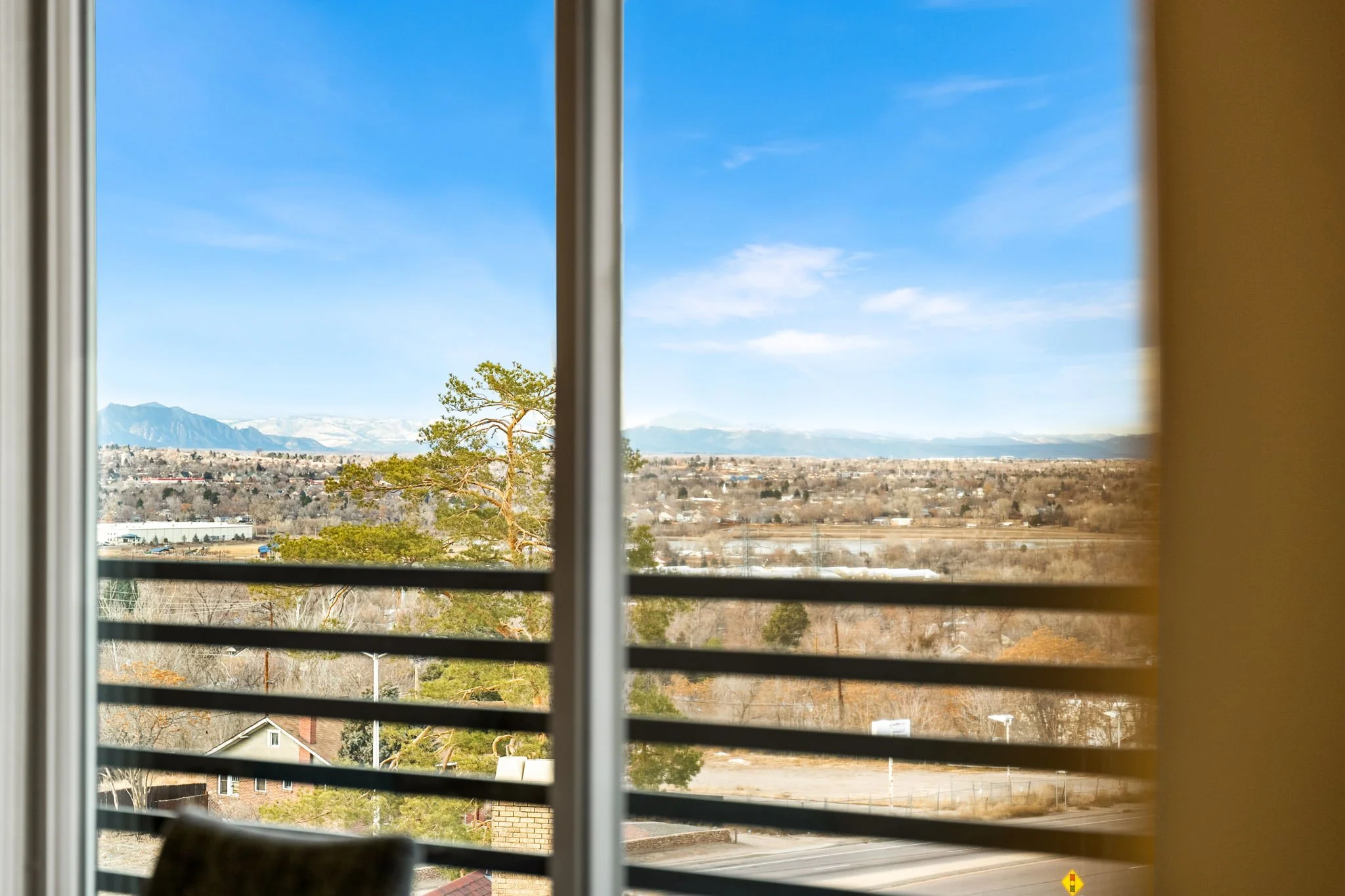 A view of a landscape with mountains, trees, and fields seen through a window with horizontal blinds.