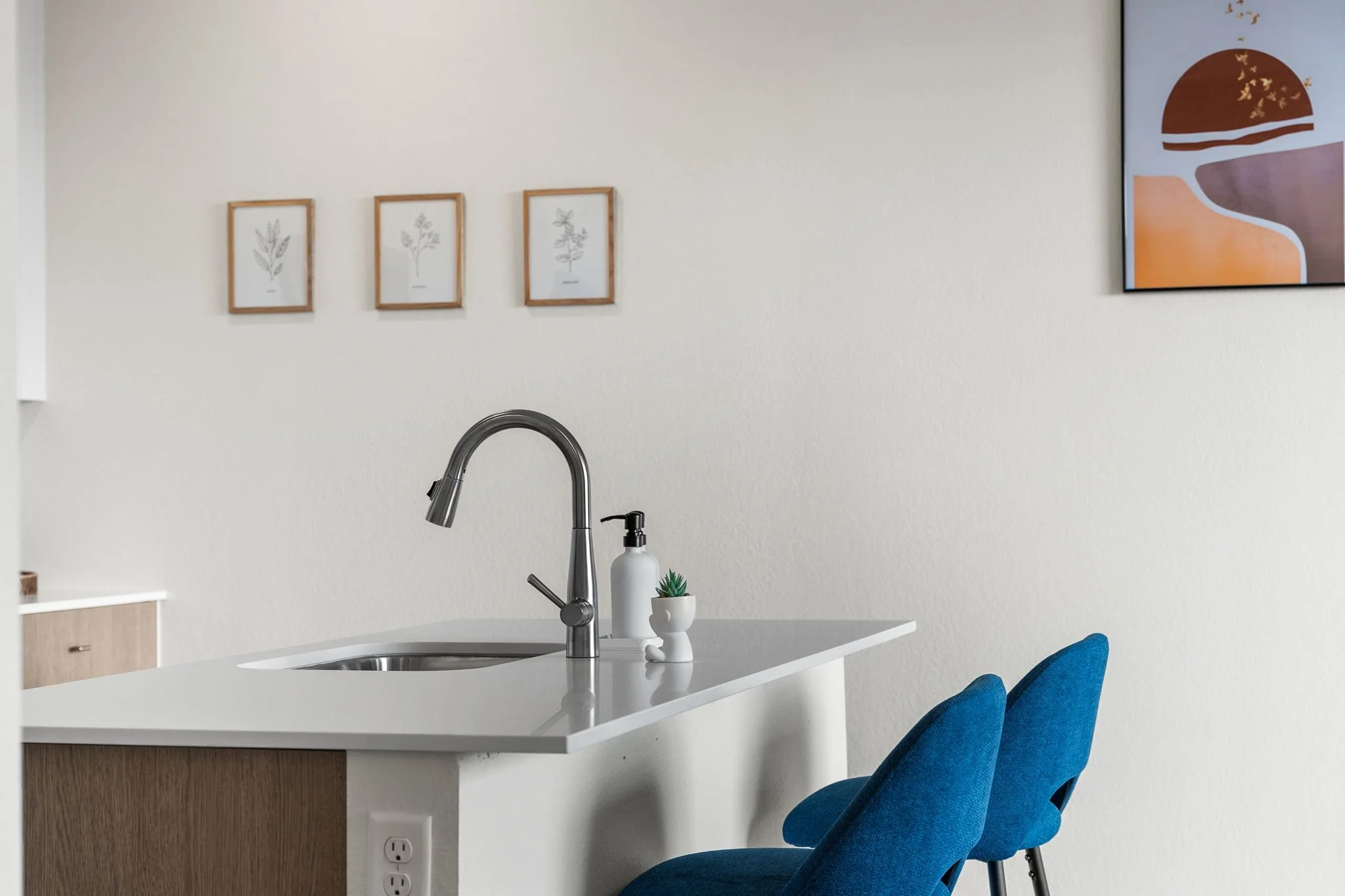 Kitchen island with a modern chrome faucet, white countertop, soap dispenser, and small potted plant. Two blue chairs are positioned at the island. Three framed botanical prints hang on the wall behind, along with a colorful abstract painting.