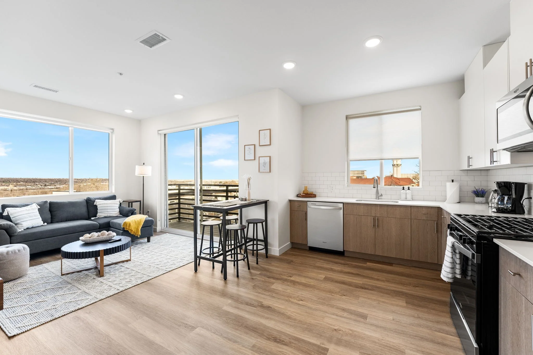 Living room and kitchen with large windows, gray sofa, fireplace, dining table with stools, kitchen cabinets, white countertops, and stainless steel appliances.