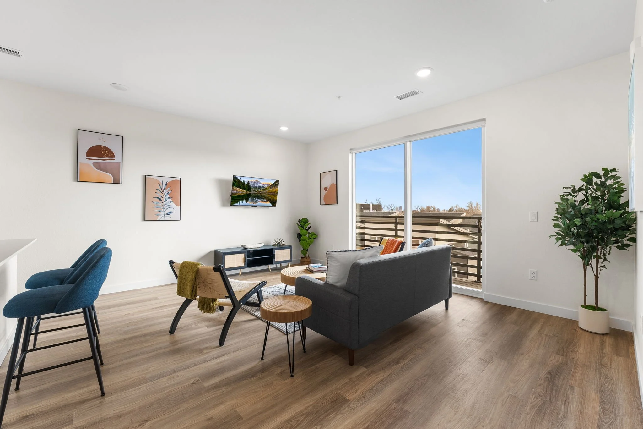 Living room with white walls, wooden floor, blue chairs, gray sofa, wall-mounted TV, colorful abstract wall art, large window with balcony, potted plant, and modern decor.