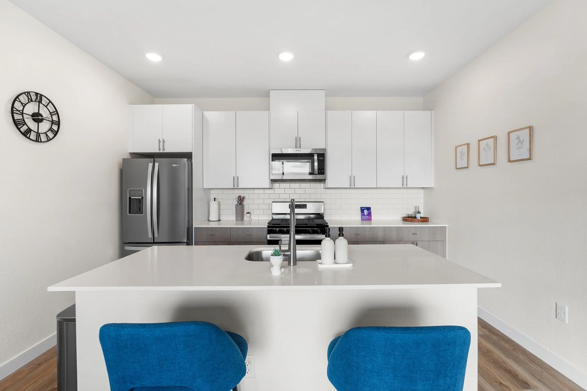 Modern kitchen with white cabinets, stainless steel refrigerator and microwave, and a white island with blue chairs.