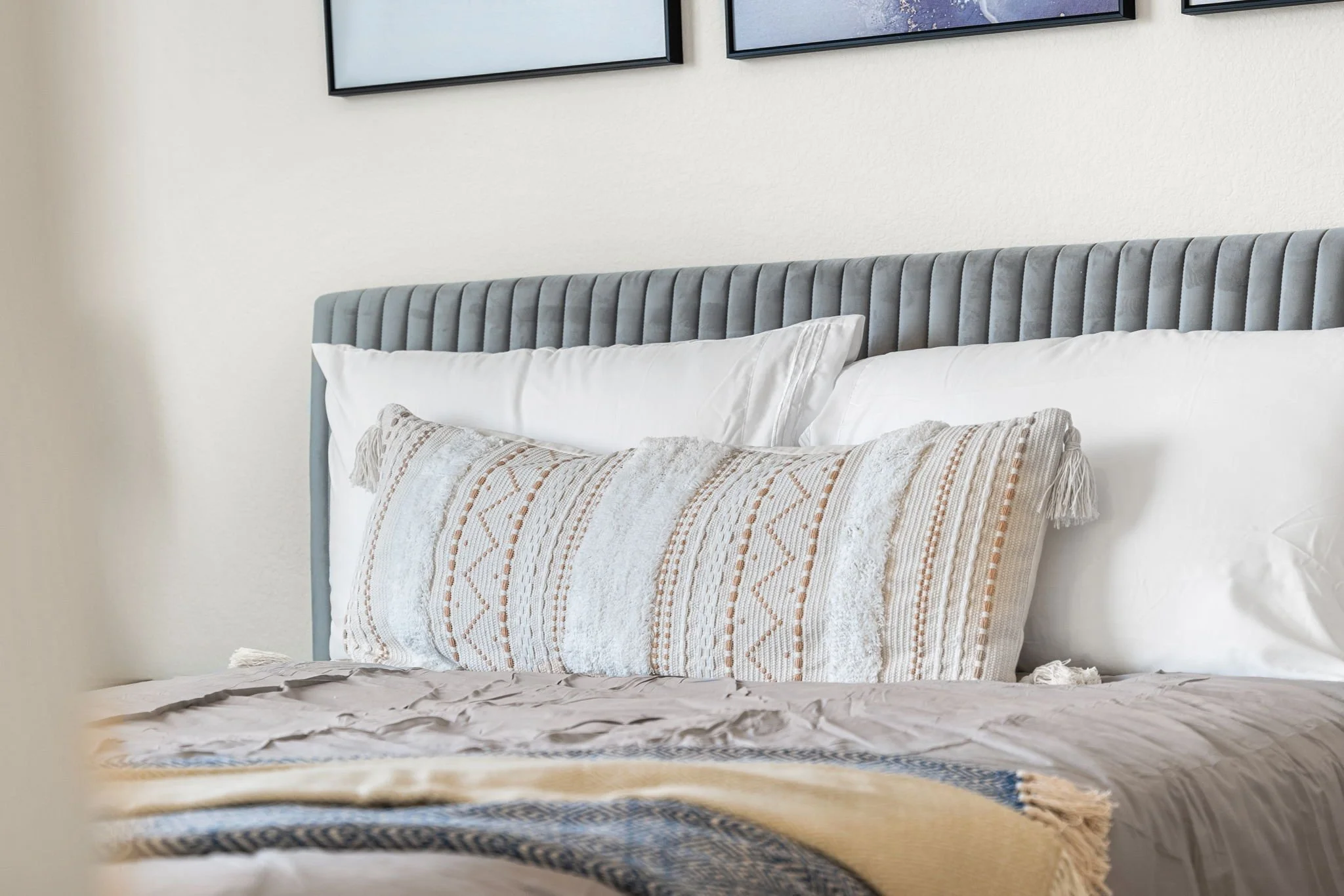 A neatly made bed with a light gray upholstered headboard, white pillows, and a decorative throw pillow, with a beige and blue blanket at the foot of the bed.