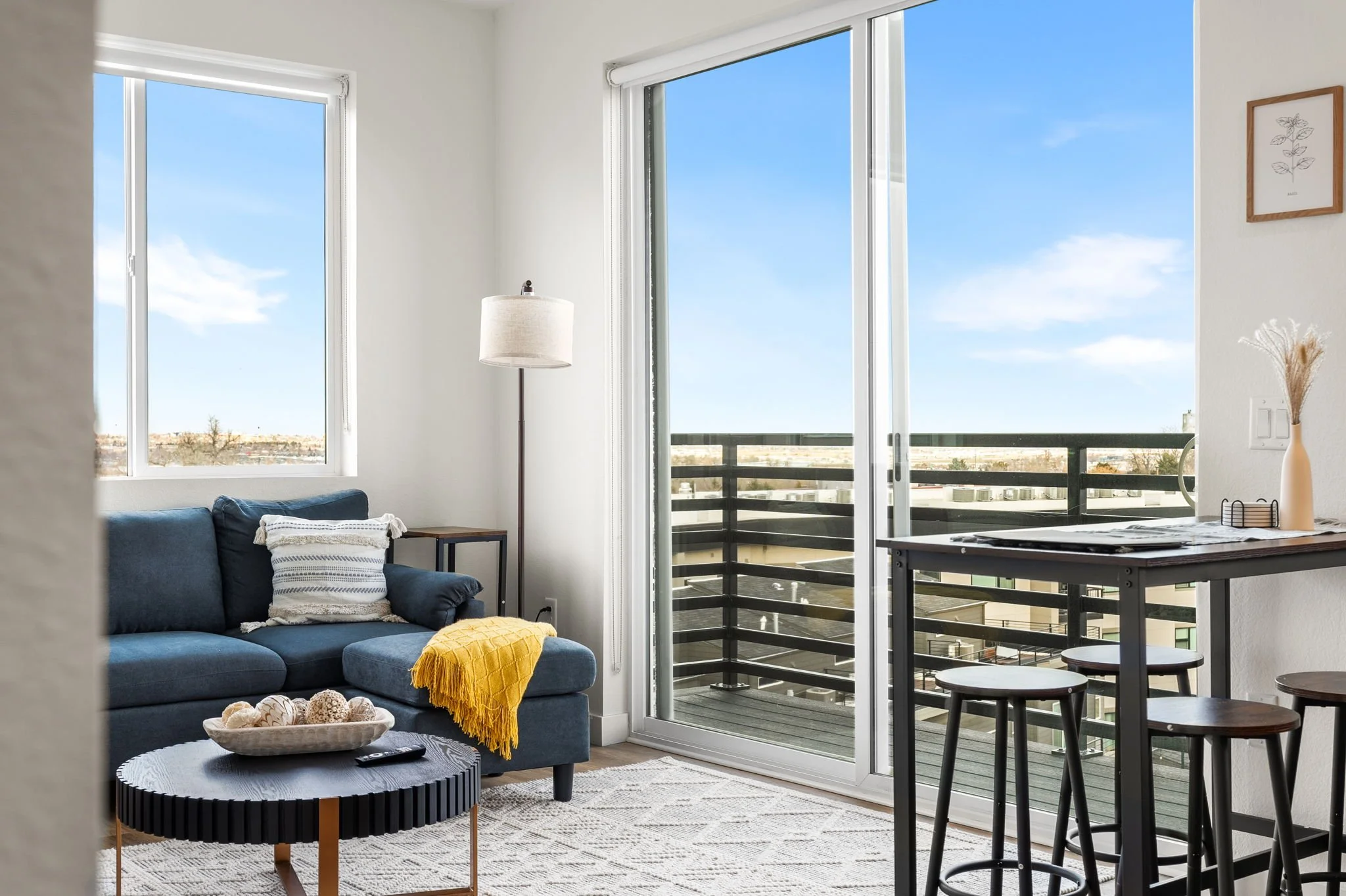 Living room with large sliding glass door leading to balcony, blue sofa with white and gray throw pillows, yellow throw blanket, black coffee table with decorative balls, floor lamp, and small table with a vase and framed wall art, balcony railing vi