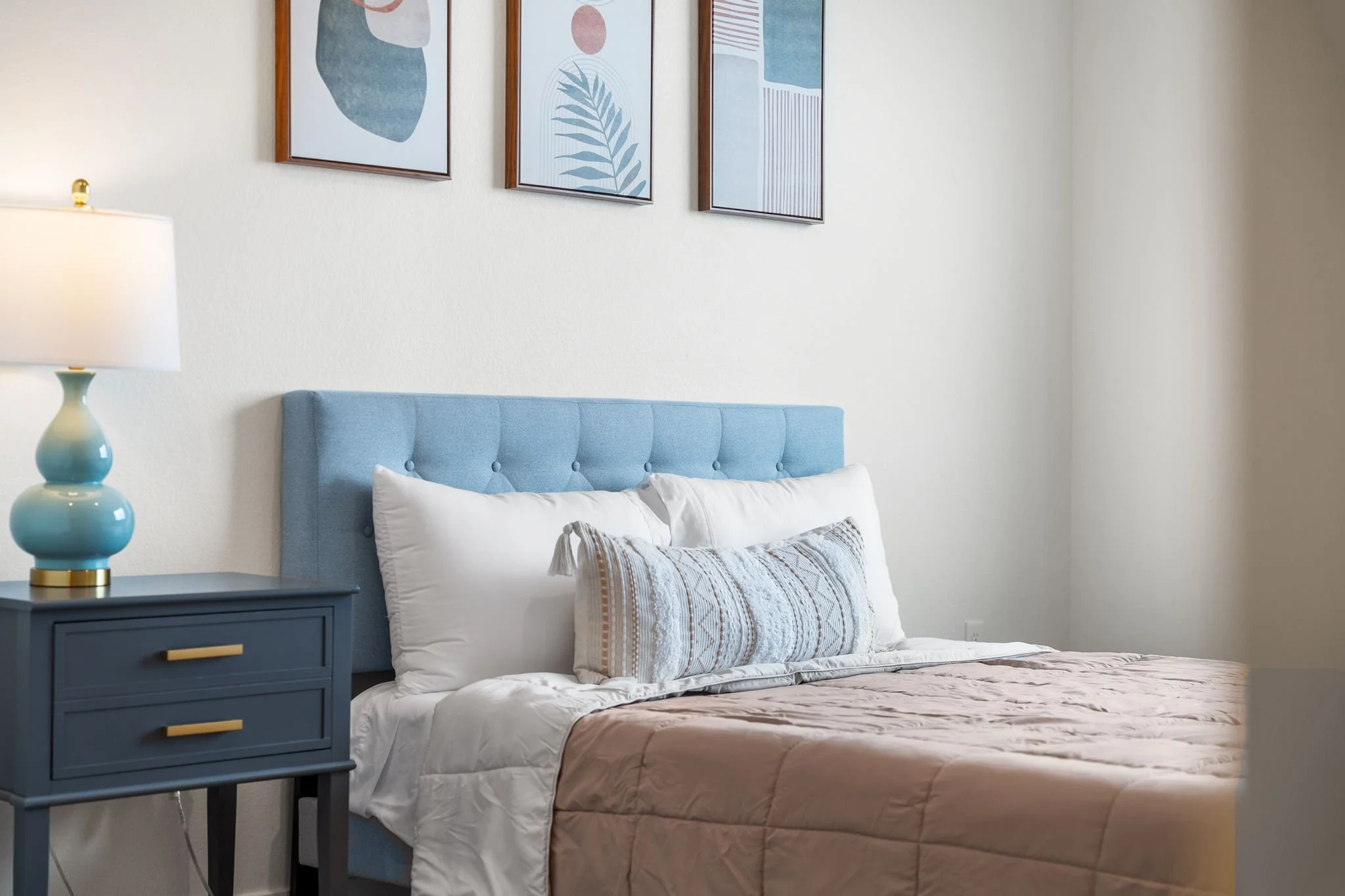 A bedroom with a light blue upholstered headboard, white bedding, and a decorative pillow. There's a blue nightstand with a blue ceramic lamp and gold accents. Three framed abstract art pieces hang on the wall above the bed.