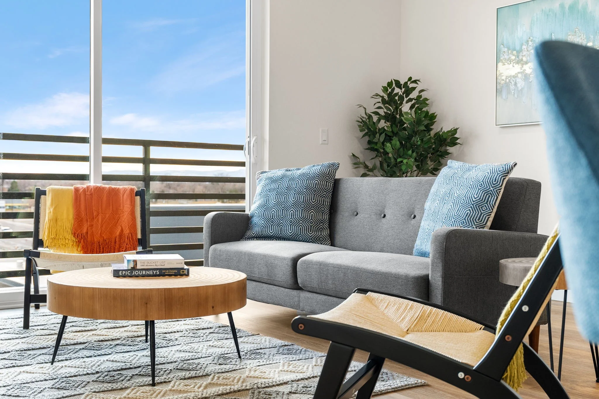 Living room with a gray couch, blue patterned pillows, a wooden coffee table with books, a black chair with an orange and yellow blanket, a sliding glass door leading to a balcony with a view of the sky, a green plant, a blue and white painting on th