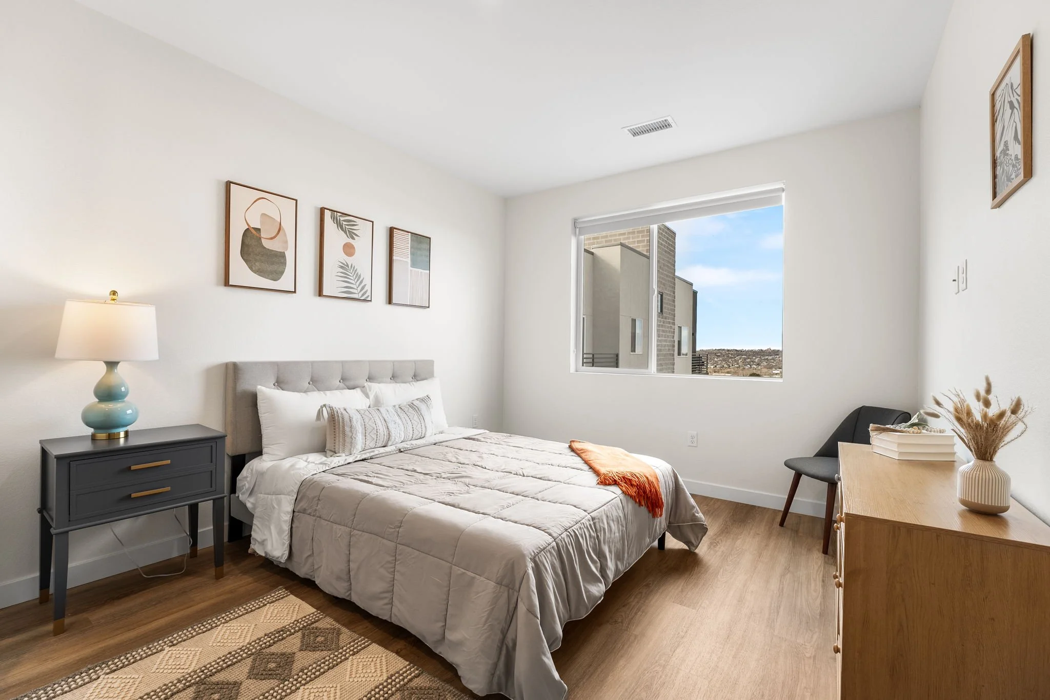 A bright bedroom with a light gray bed, white bedding, a black nightstand with a blue lamp, a window showing a cityscape, abstract wall art, and a wooden dresser with a vase and books.