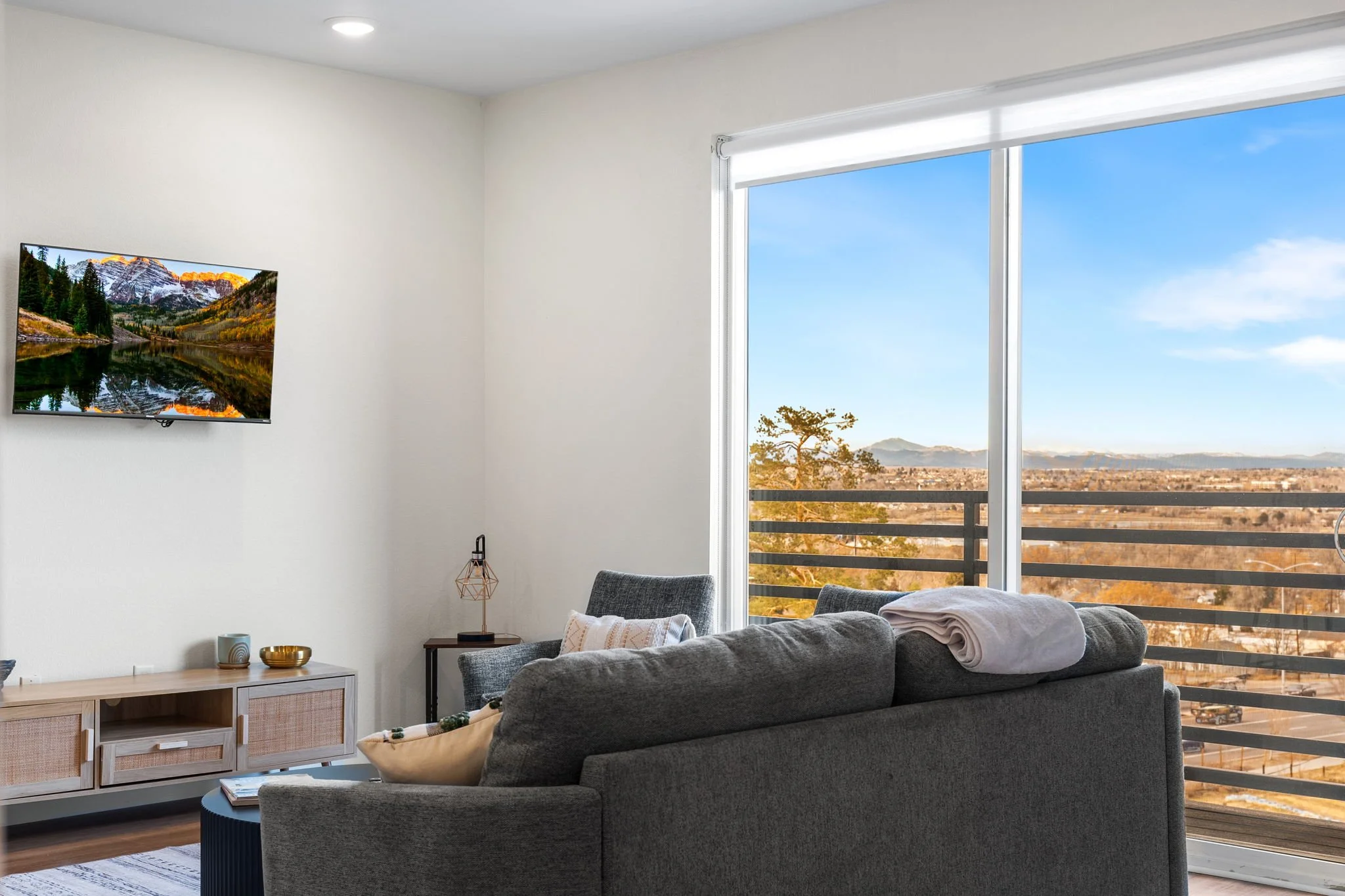 Modern living room with large sliding glass door opening to a balcony with a view of mountains, a television mounted on the wall, and contemporary furniture including a gray sofa and chairs.