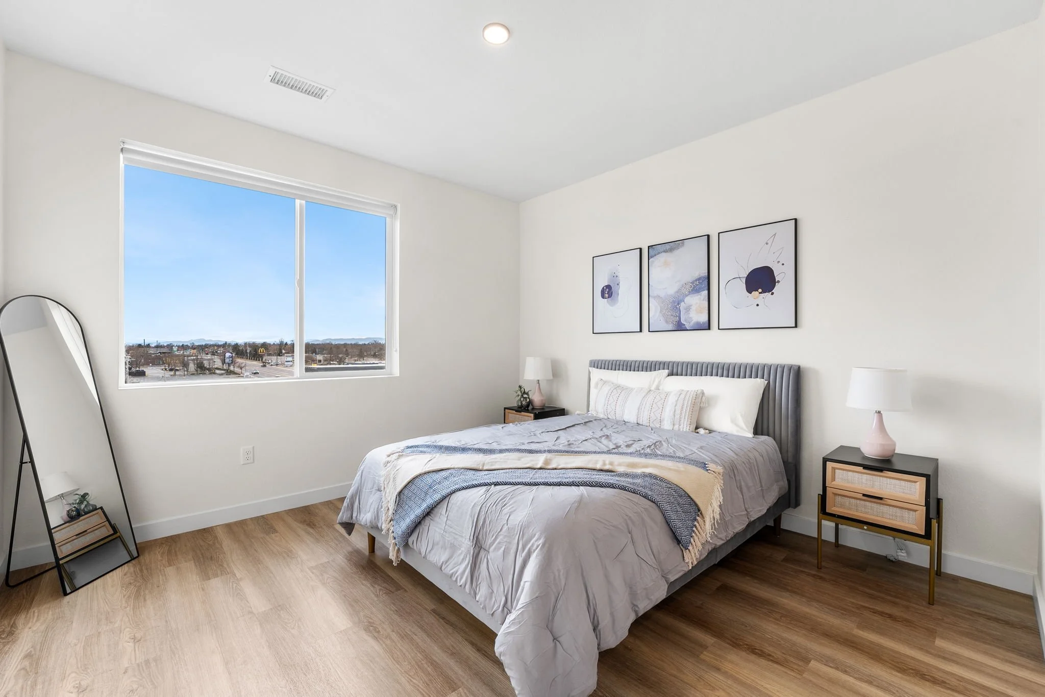 A bedroom with a large window showing a clear sky and distant mountains. The room has a bed with a gray headboard and white bedding, flanked by two black nightstands with pink and white lamps. A tall mirror leans against the wall on the left. The wal