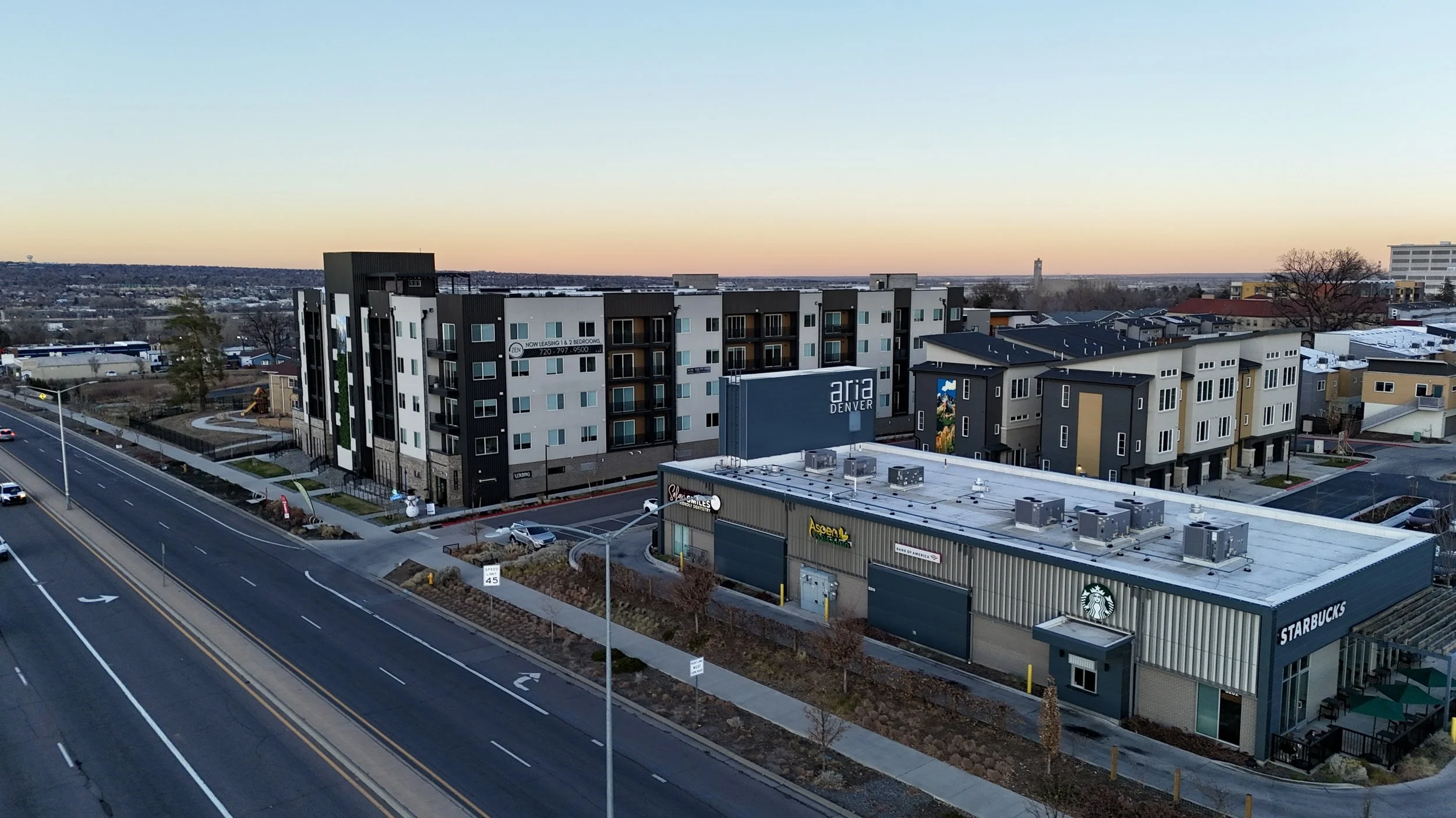 Urban scene at sunset showing a parking lot, a Starbucks cafe with outdoor seating, a mixed-use building with apartments above, and signs for Aria Denver and other businesses.