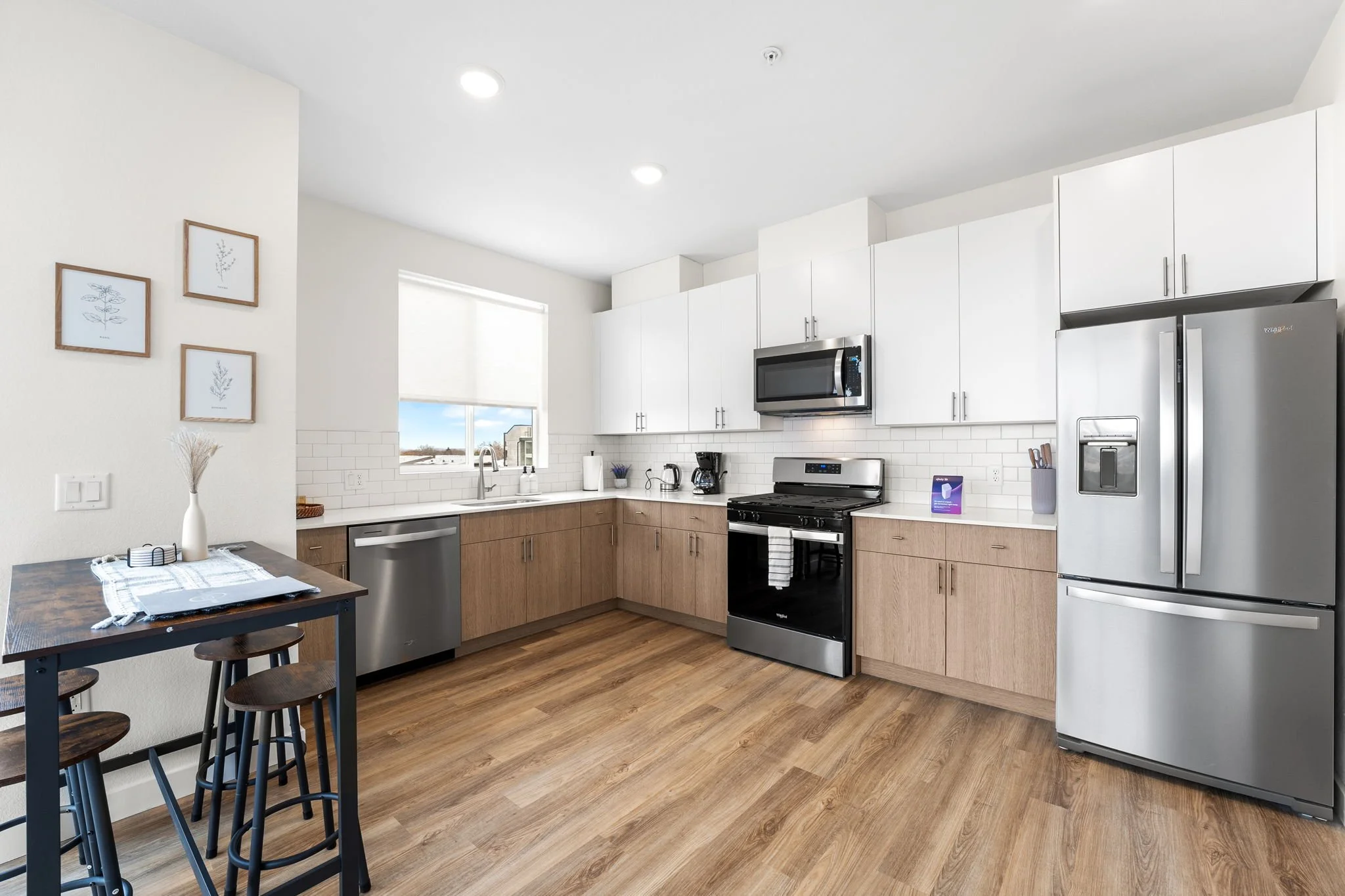Modern kitchen with white upper cabinets, wood lower cabinets, stainless steel appliances including refrigerator, oven, microwave, and dishwasher, white subway tile backsplash, and wood flooring. A small dining table with three black stools is on the