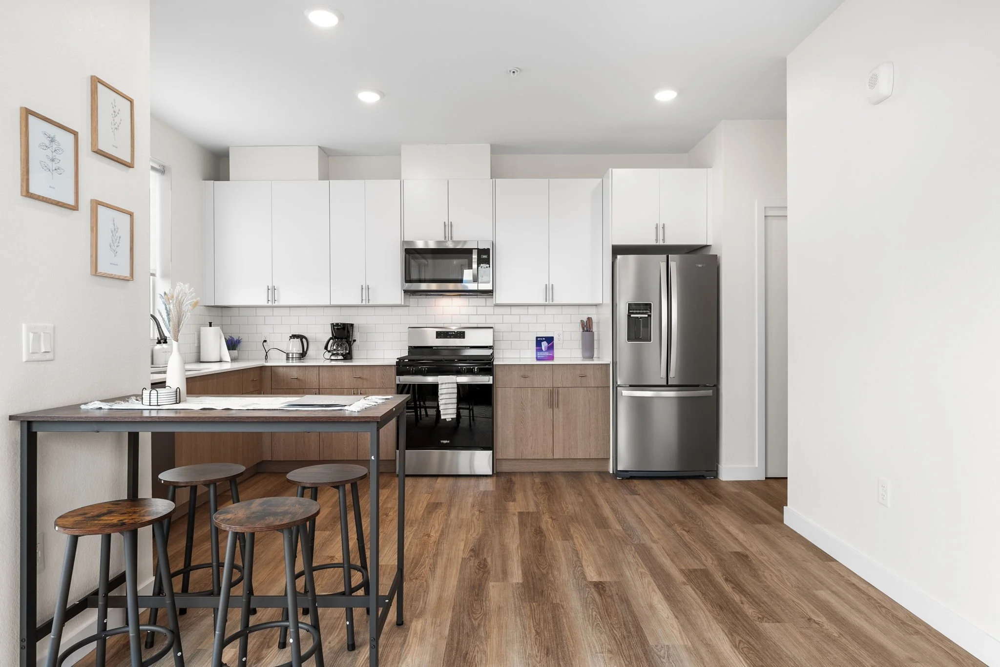 Modern kitchen with white cabinets, stainless steel appliances, a small dining table with three stools, wooden flooring, and framed botanical artwork on the wall.