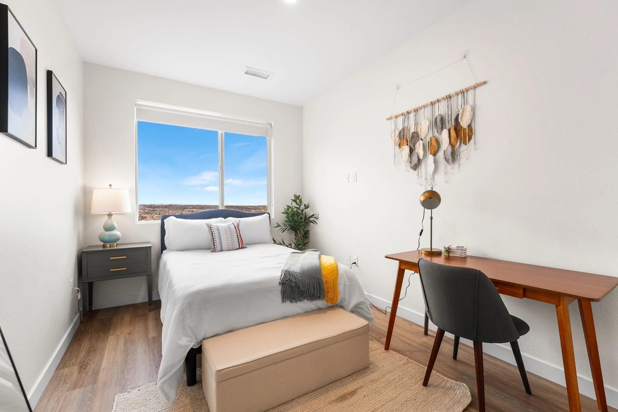 A bedroom with a bed, nightstand, lamp, window, desk, chair, wall art, and decorative wall hanging.