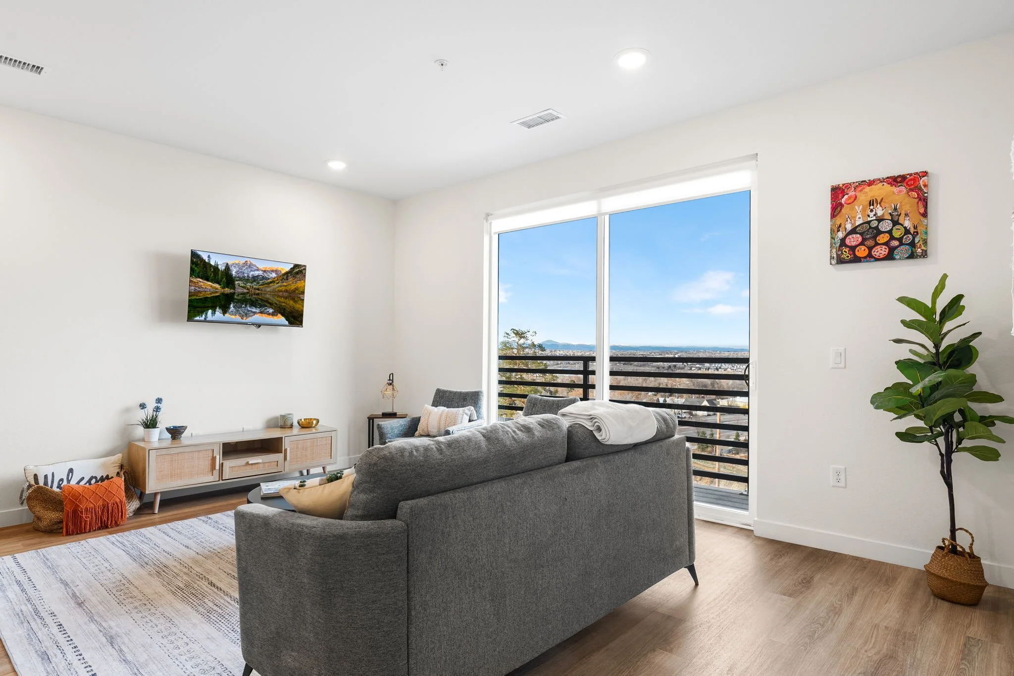 Living room with gray sofa, large window with balcony view of cityscape and distant mountains, wall-mounted TV, woven accent pillows, potted plant, colorful artwork, wooden floor, white walls, ceiling lights.