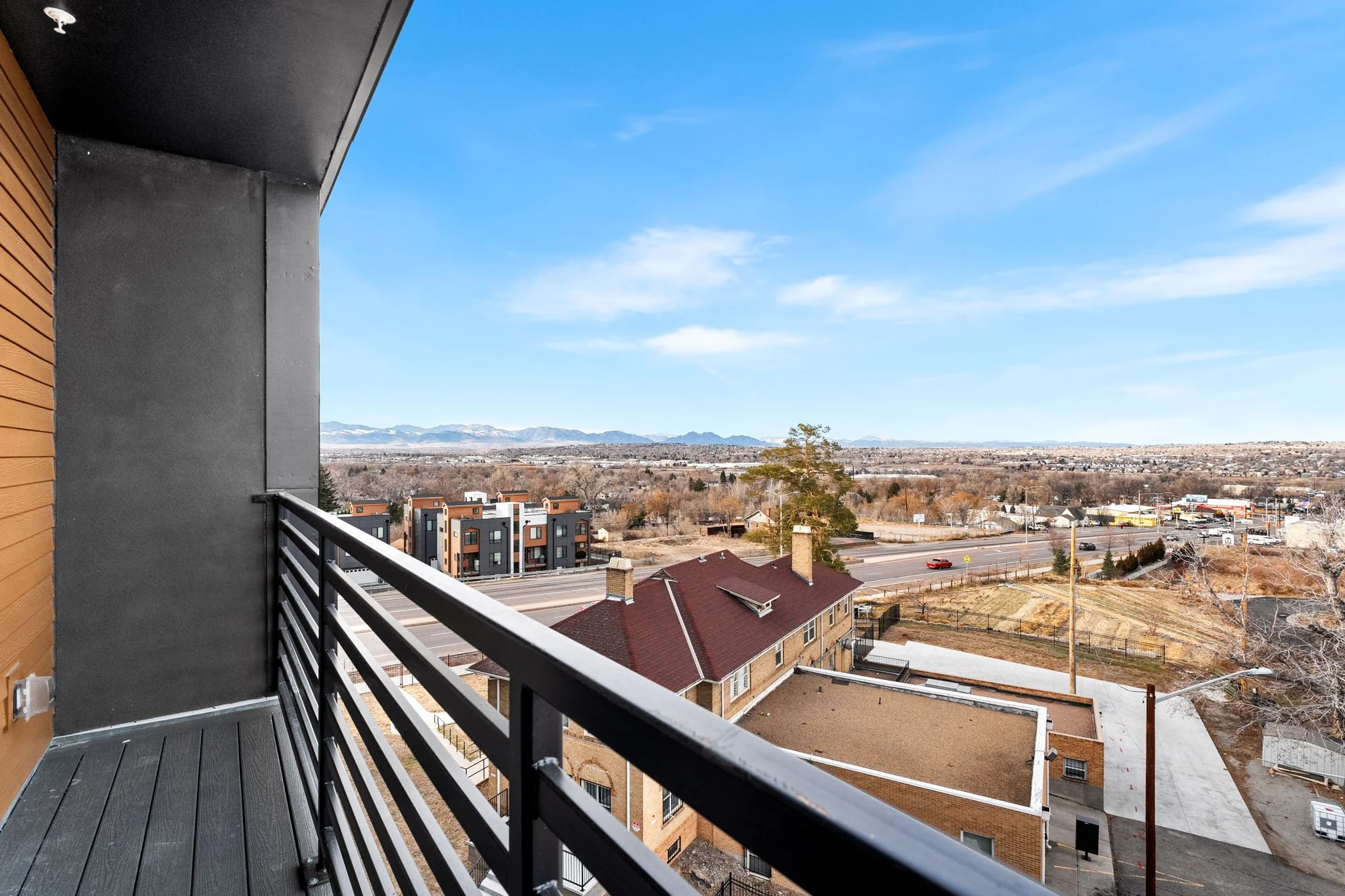 View from a balcony overlooking a residential area with houses, a road, and mountains in the distance under a blue sky.
