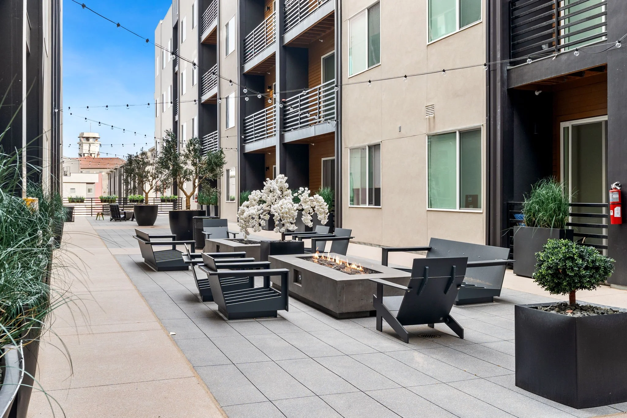 Outdoor patio area with black chairs and a rectangular fire pit, large white floral arrangement, potted plants, and string lights on a multi-story residential building with balconies.