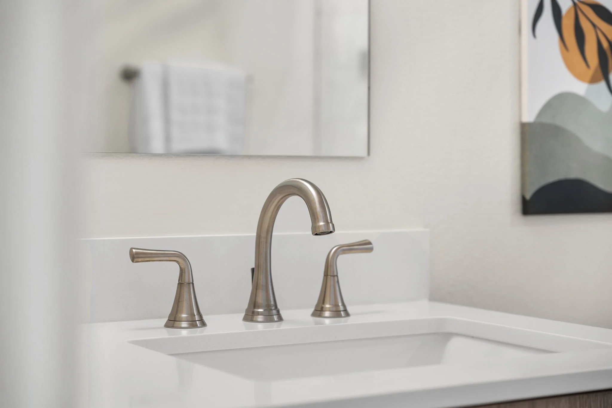 Bathroom sink with brushed nickel faucet and two handles, a mirror above, and a piece of abstract wall art on the wall to the right.