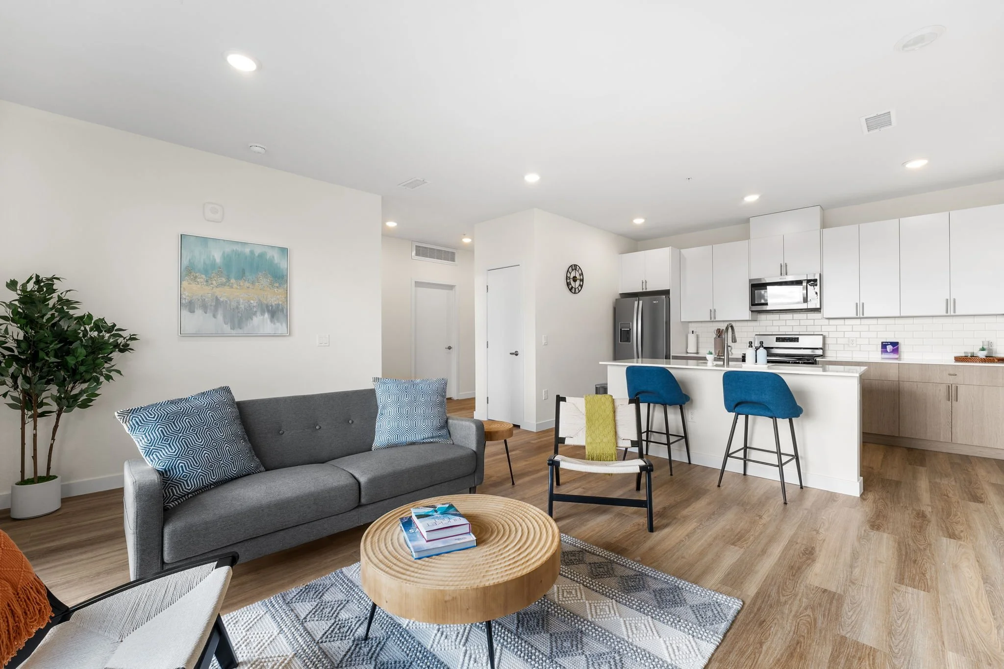 Open-concept living room and kitchen with gray sofa, wooden coffee table, blue bar stools, white cabinets, stainless steel appliances, and hardwood floors.