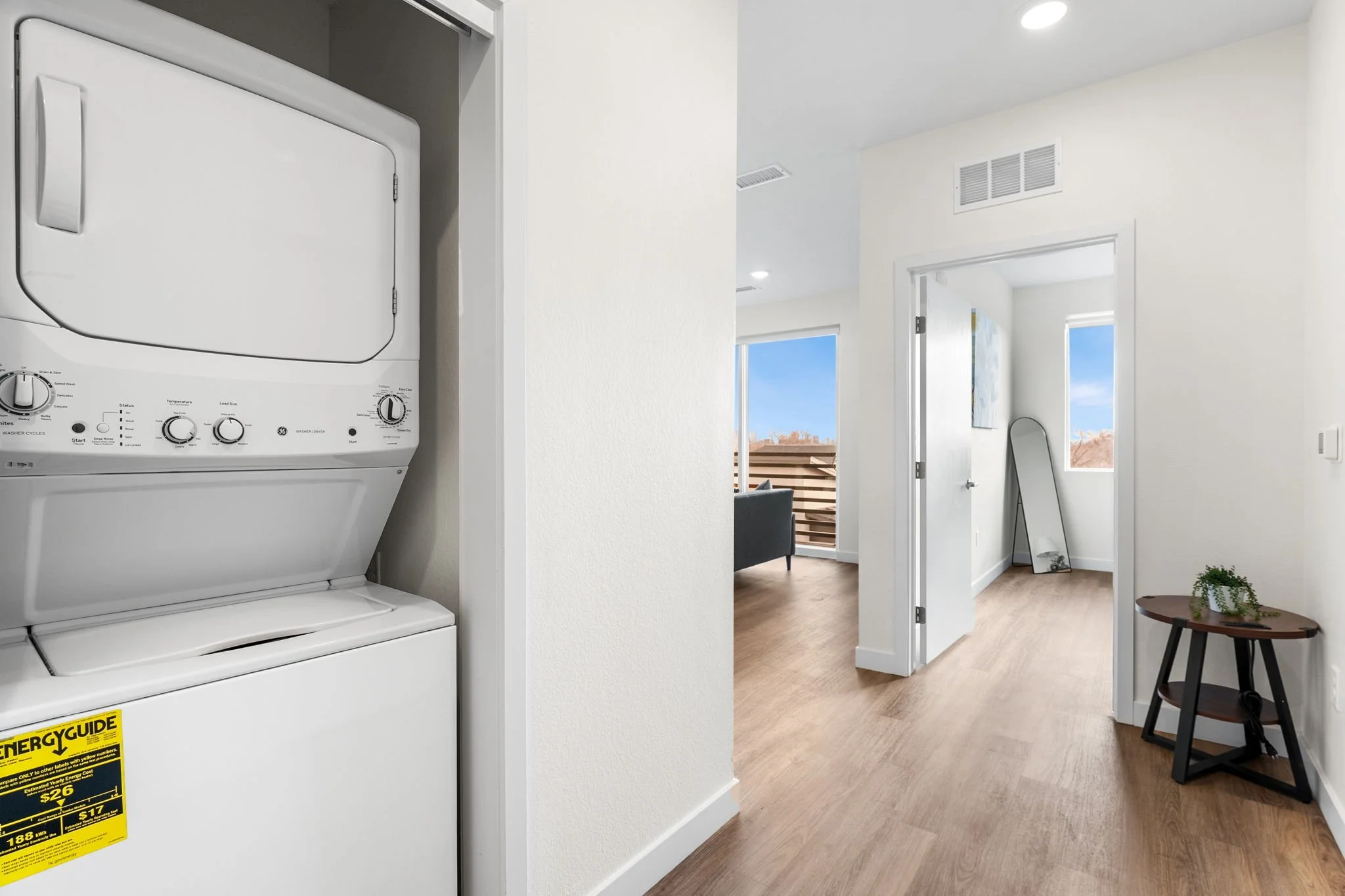 Laundry area with a stacked washer and dryer to the left, and a hallway leading to a living room and another room in the background with windows and wooden flooring.