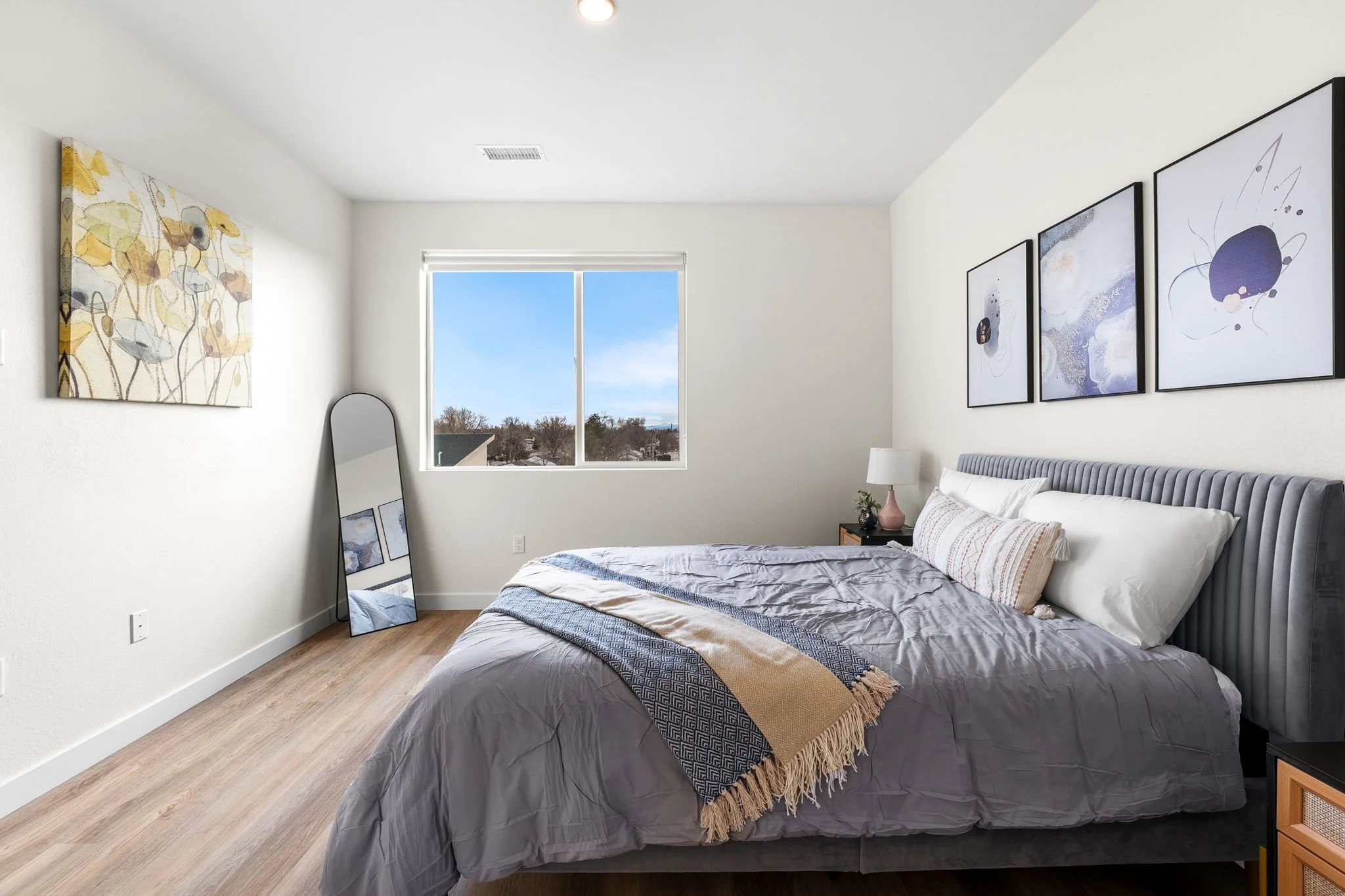 A bedroom with a bed that has a gray headboard, white pillows, and a blanket. There is a window with a view of the outside, white walls with abstract artwork, a mirror leaning against the wall, a nightstand with a lamp, and wooden flooring.