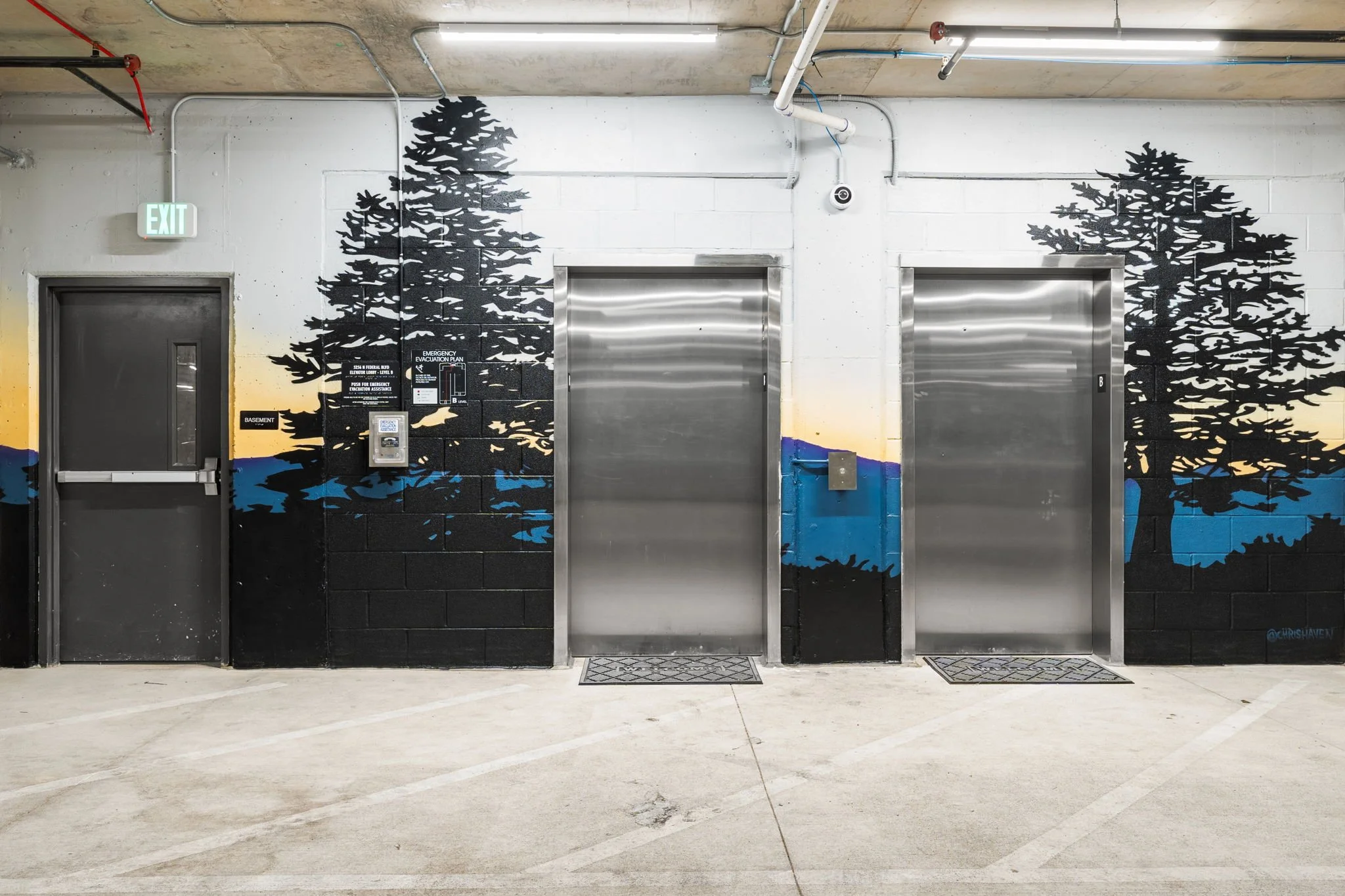 Street art mural of trees on a concrete wall with two elevators and a door in a parking garage.