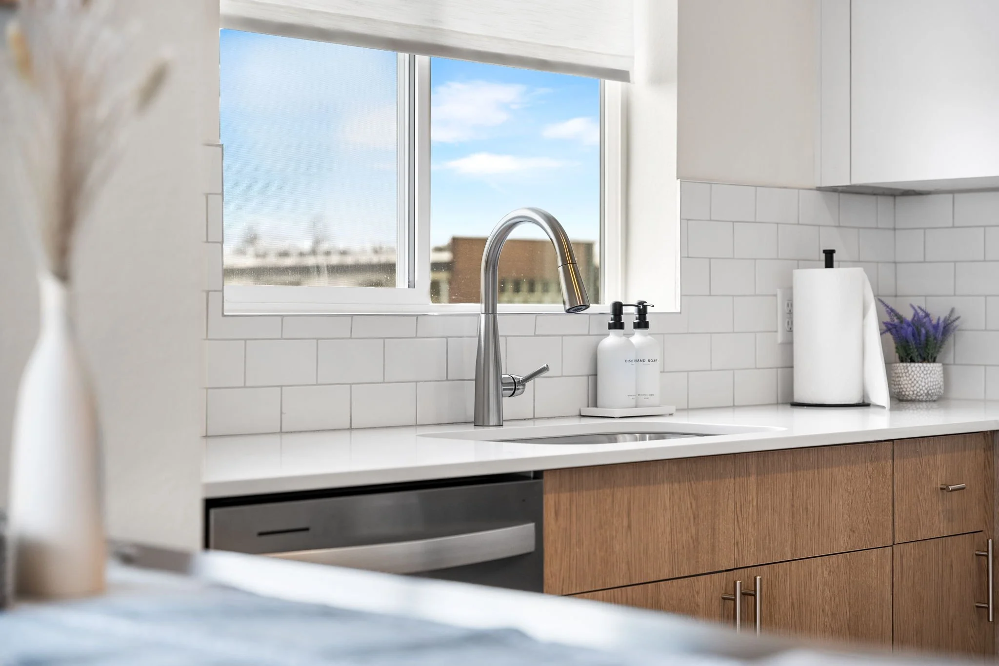Modern kitchen with white countertops, a window with blue sky view, a stainless steel faucet, soap dispensers, a paper towel holder, and a small plant on the counter.