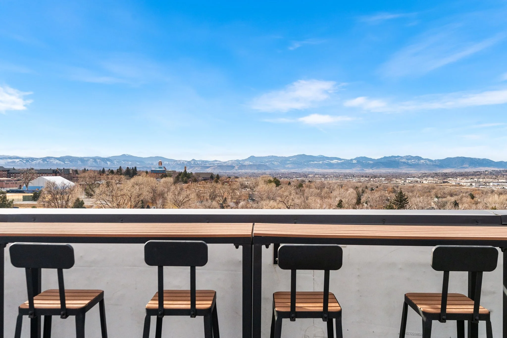 Patio with four wooden and black chairs, overlooking a cityscape and distant mountains on a sunny day.