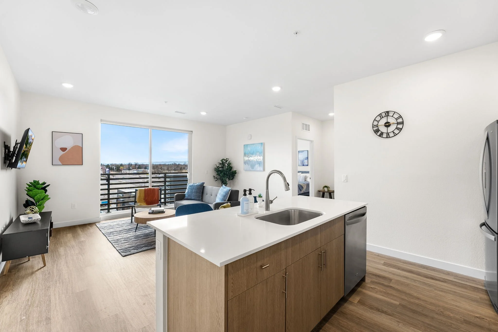 Open living room with sliding glass door leading to balcony, modern furniture, artwork on walls, and kitchen island with sink and dishwasher.