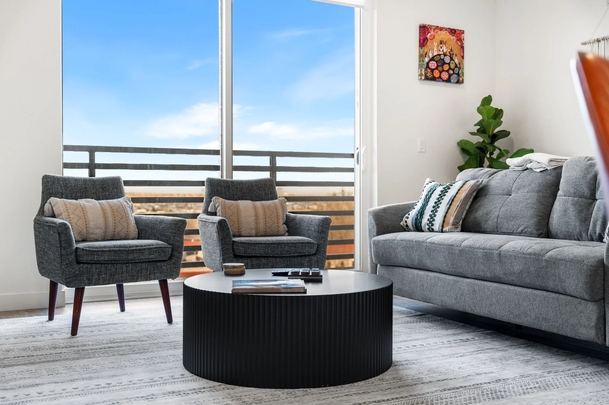 Modern living room with gray sofa and two gray armchairs, a black round coffee table with books and remote, a large sliding glass door with balcony view of blue sky, a colorful wall art, a green plant, and a white and gray patterned rug.