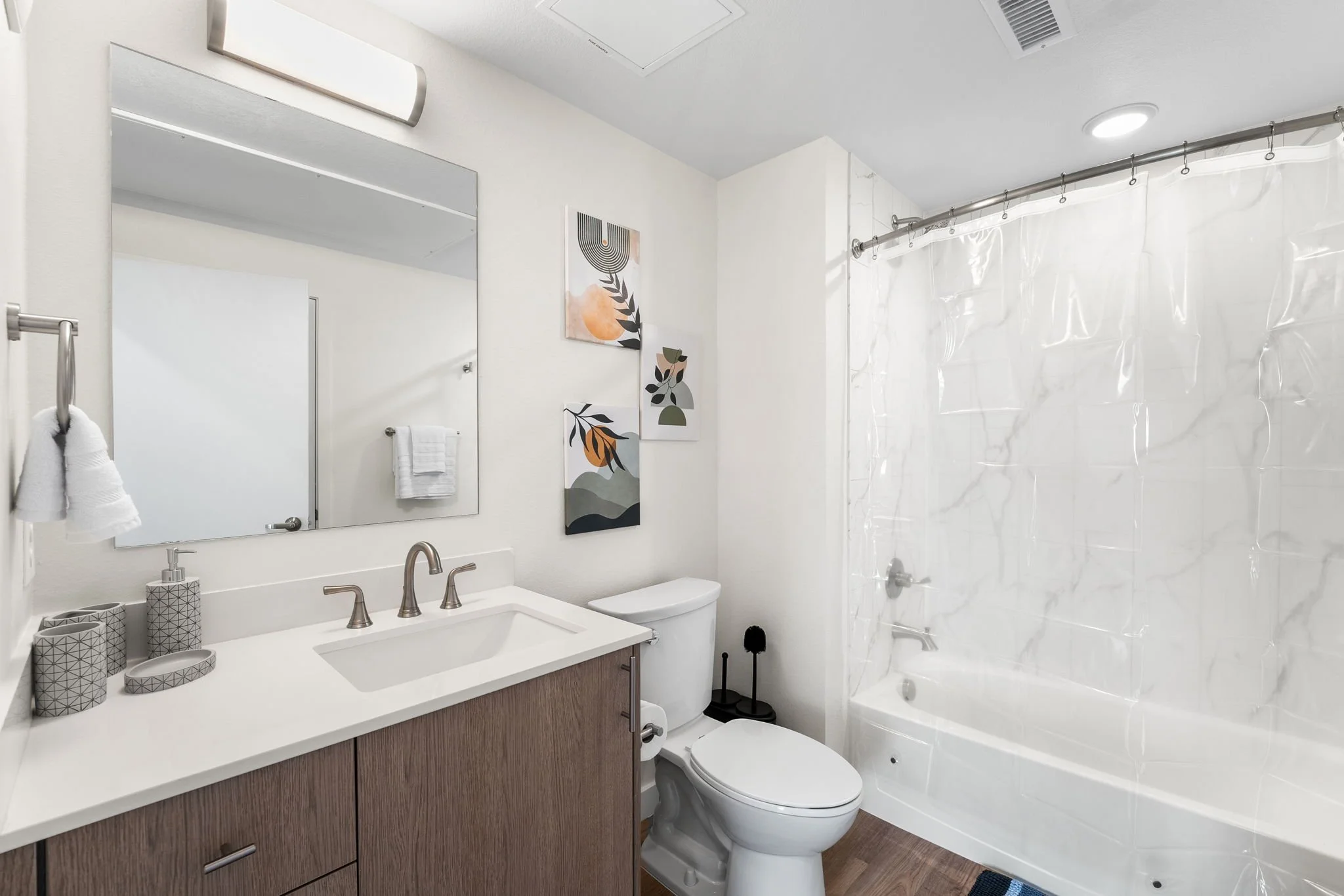 Modern bathroom with white walls, a large mirror, a wooden vanity with a white countertop, a single sink, wall-mounted faucet, decorative containers, a toilet, abstract art on the wall, a shower with a clear plastic curtain, and wooden flooring.