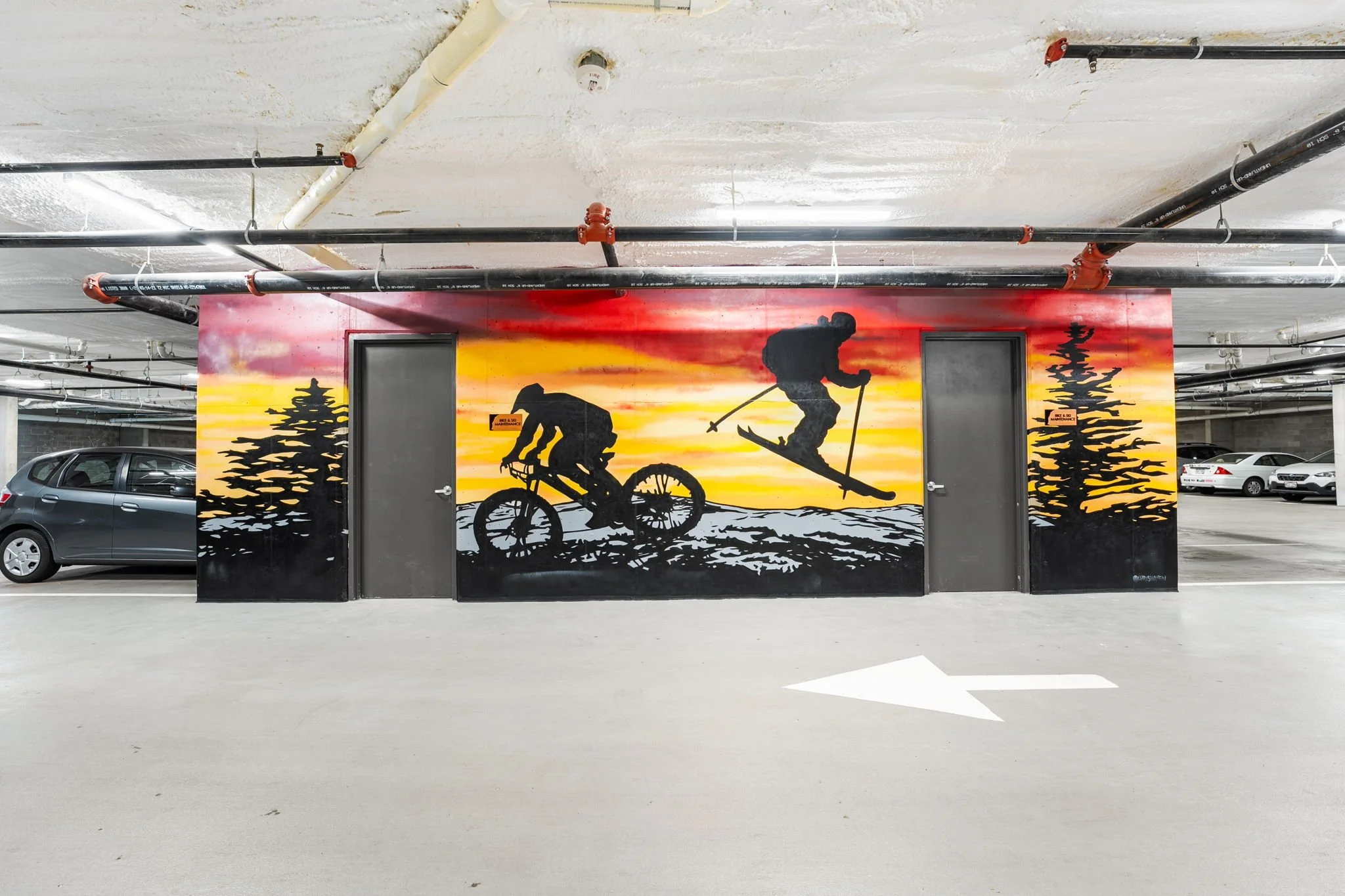 Mural of silhouettes of a person mountain biking and a person snowboarding with a sunset background and pine trees, painted on a wall in an underground parking garage.